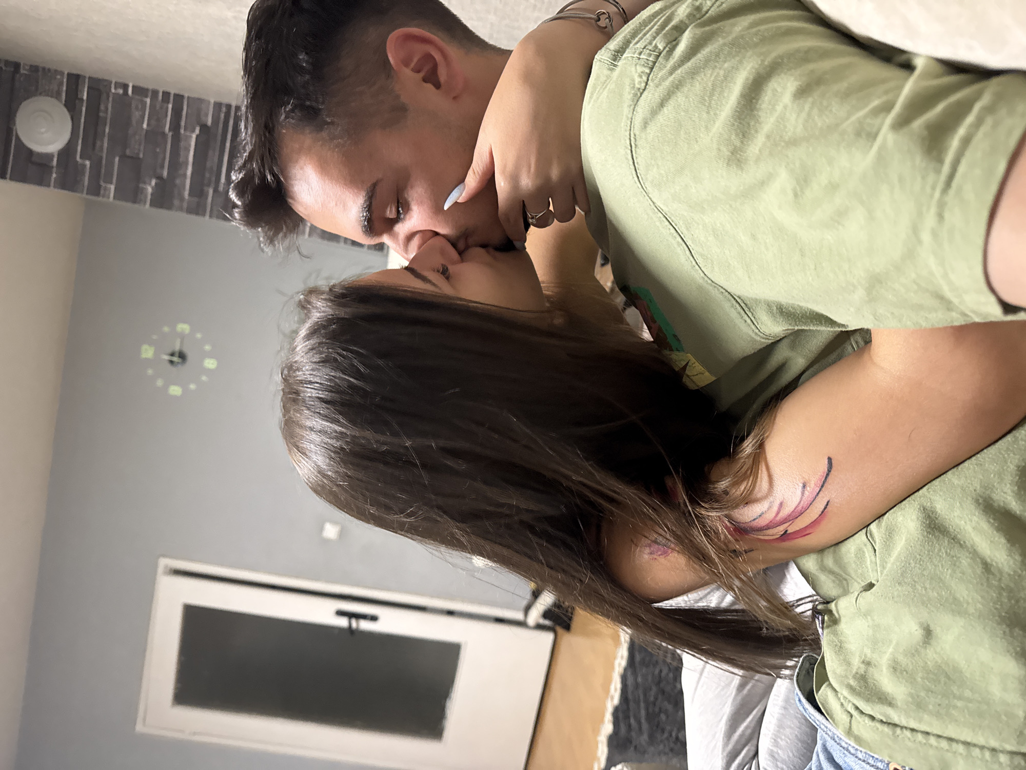 The image captures an intimate moment between two young individuals, presumably a couple, sharing a passionate kiss. They are depicted from an overhead, slightly tilted perspective, suggesting they are reclining indoors.
The female subject, positioned on the right, has long dark brown hair and is wearing a light green short-sleeved top and light blue jeans. Her left arm, adorned with an abstract tattoo of red and black curved lines on the bicep, is wrapped around the male's neck. Her hand, featuring long light blue painted fingernails and a silver ring on her ring finger, gently touches his jawline.
The male subject, on the left, has short dark hair and is wearing a dark garment. A silver bracelet is visible on his left wrist. Both individuals have their eyes closed, conveying deep affection and tenderness during the kiss.
The indoor setting, potentially a home in Sofia, Bulgaria, features a textured grey wall behind the male's head, topped by a white circular smoke detector or similar device. A light grey wall is visible with a round, illuminated clock displaying green numbers and hands, possibly indicating a time around 11:15 or 12:15. A white-framed window or similar fixture with a dark pane is present in the lower left background. The overall atmosphere is one of closeness and personal affection. No legible text is visible in the image.
