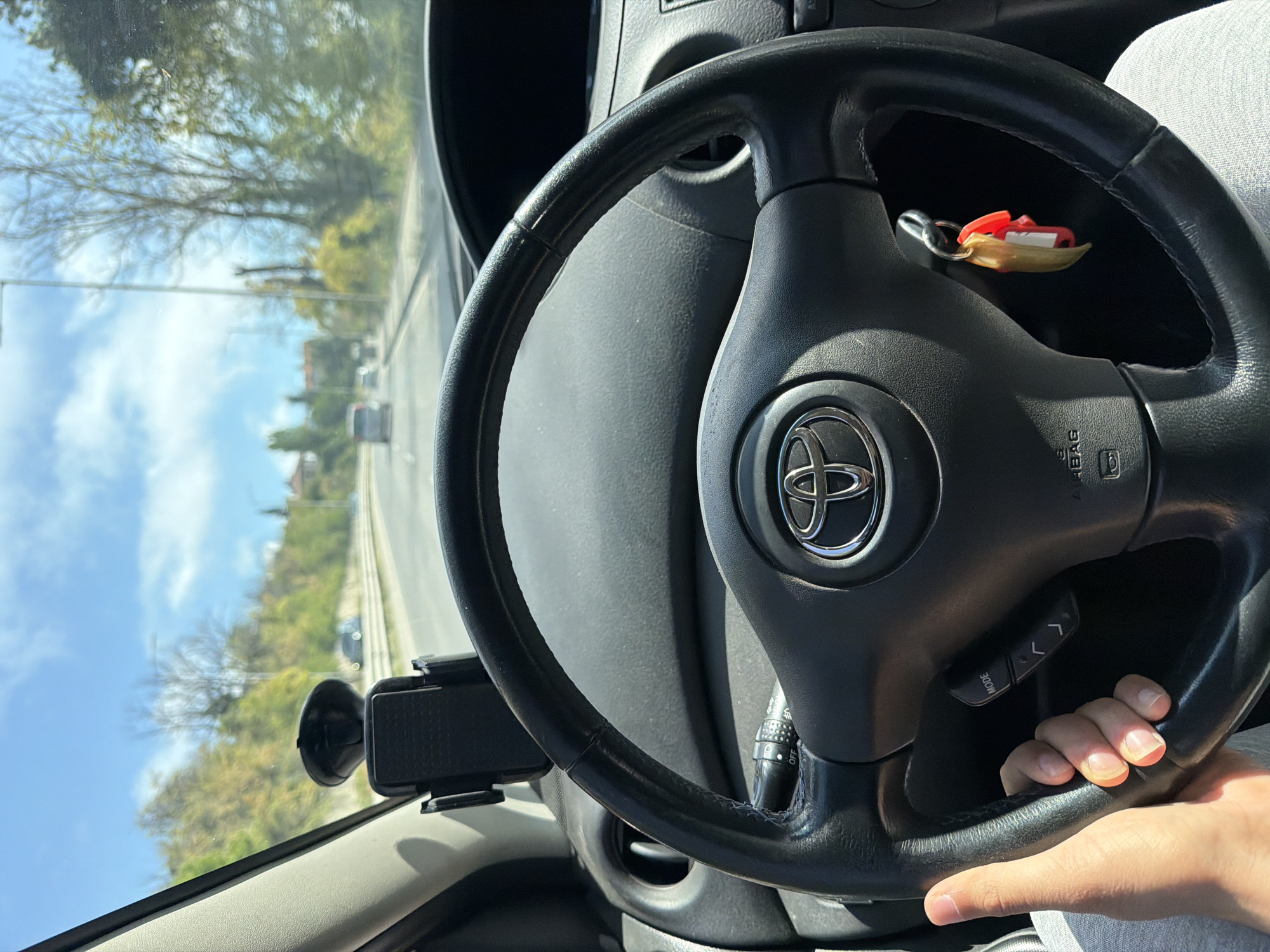 The image shows the interior of a car, likely a Toyota, from the driver's perspective. The steering wheel is prominently displayed, with the Toyota logo visible in the center. Keys are hanging from the steering wheel. The driver's left hand is on the steering wheel, near the mode controls. A phone holder is visible on the left side of the dashboard. The view through the windshield shows a road with other cars, trees, and a blue sky. Based on the architecture and vegetation, this is likely Varna, Bulgaria.