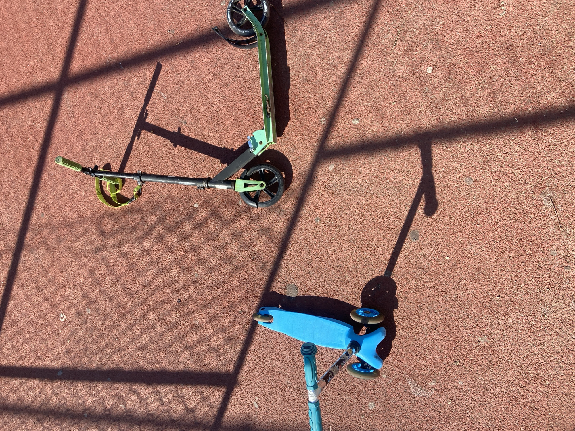 Two scooters are on a red textured surface. One scooter is green and black, lying horizontally. The other is blue and partially visible in the lower right corner. Shadows from an unseen object are cast across the surface. The location is unspecified.