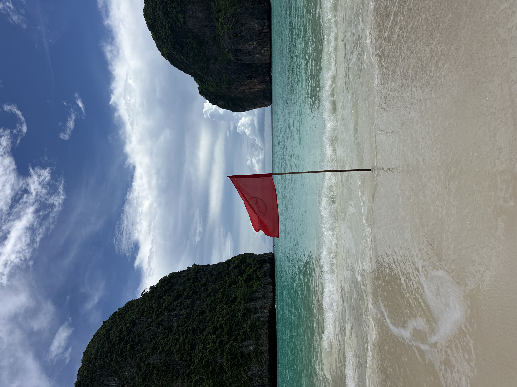 A red flag with a circular symbol, possibly indicating a warning or restriction, is planted in the sand on a beach. The flag is positioned centrally in the foreground, slightly off-center to the left. Behind the flag, turquoise water with gentle waves laps at the shore. Two large, green, rocky cliffs flank the beach, one on the left and one on the right, forming a cove-like environment. The sky above is a mix of blue sky and dramatic, gray and white clouds. The scene is consistent with the scenic beaches and karst landscapes found in the Ao Nang area of Krabi province, Thailand, known for its stunning natural beauty and island hopping tours. There are no people visible in the image.