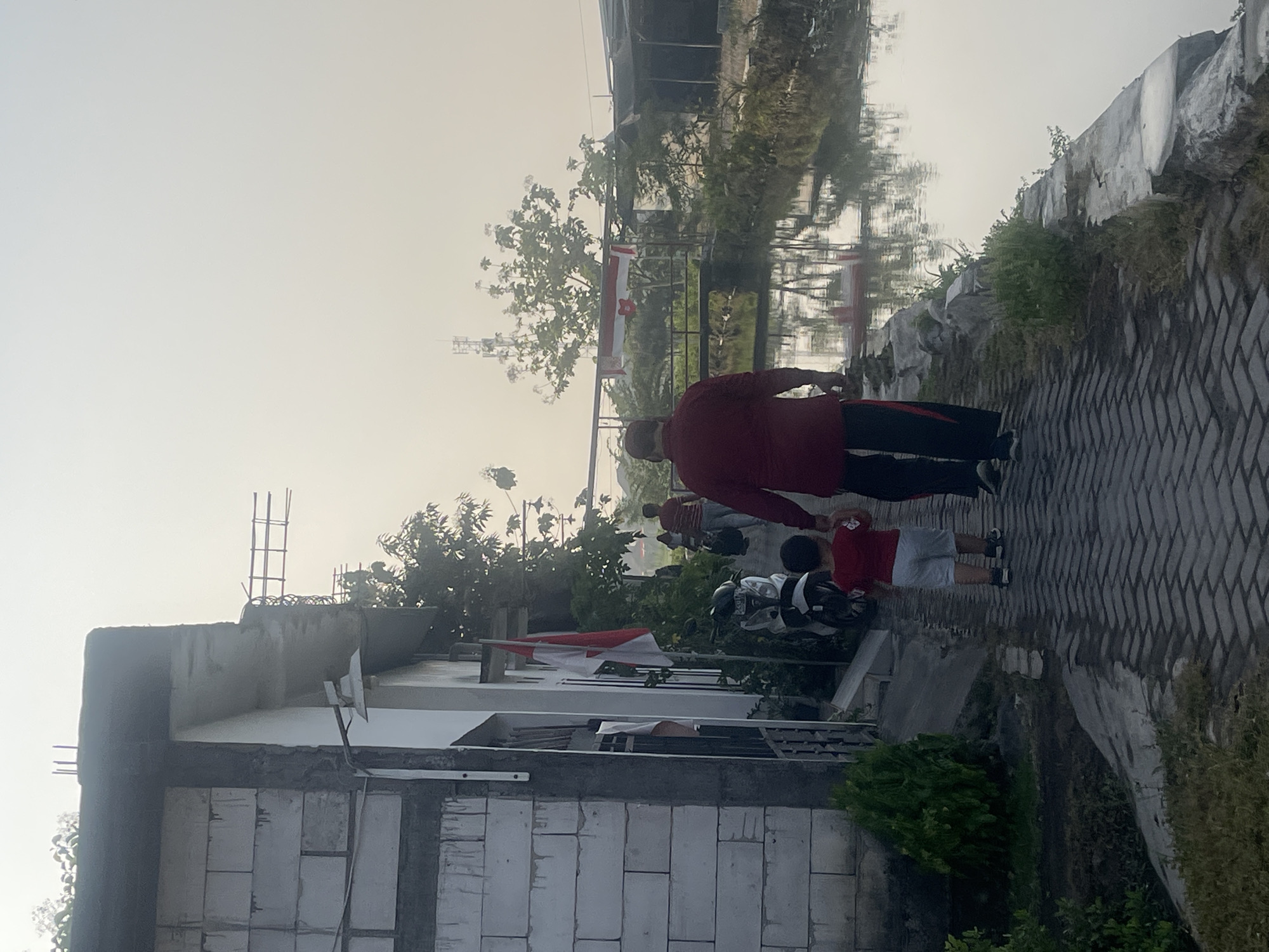 In Sugih Waras, Indonesia, a man in a red shirt and dark pants, possibly an adult, walks hand-in-hand with a young child in a red shirt and white shorts on a brick pathway. To their left is a white brick building with exposed rebar and concrete framing, and a motorcycle parked near it. An Indonesian flag is visible attached to the building. To their right, a calm body of water reflects the hazy sky and surrounding trees. In the background, a small bridge crosses the water, and a telecommunications tower is visible in the distance. The overall atmosphere appears serene, with a soft, diffused light suggesting it is early morning or late afternoon.