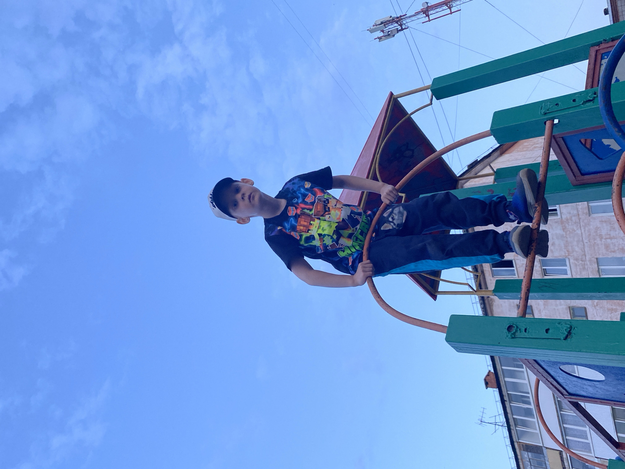 A young boy in a baseball cap and patterned t-shirt is on a piece of playground equipment in what appears to be the city of Noviy, Russia. He is holding onto circular bars, with his feet visible on the equipment. The background consists of a blue sky with some clouds, a building, and a telecommunications tower. The playground equipment is painted in green, blue, red, and brown. The photograph is taken from a low angle.