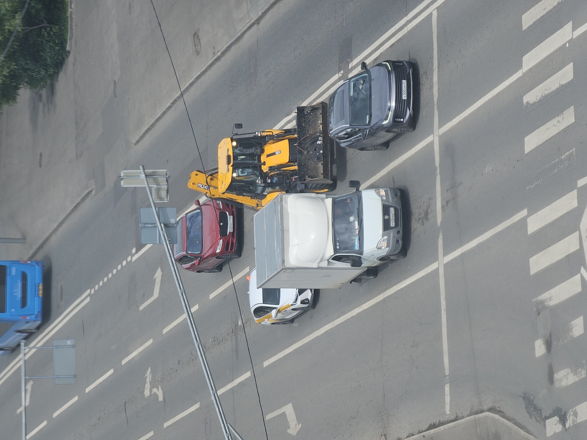 The image shows a street scene in Moscow, Russia, with several vehicles on a multi-lane road. In the foreground, a dark gray SUV with license plate X300OH37 is driving in the right lane. To its left, a white truck with a box trailer is in the center lane. Behind the truck, a white sedan with yellow and orange markings, possibly a taxi, is in the left lane. A red SUV is in the lane to the left of the taxi. A yellow backhoe loader is in the right lane, partially blocking the road. Further up the road, a blue bus is visible in the distance. The road is marked with white lane lines and a pedestrian crossing. The environment appears to be an urban setting with some trees visible on the right side of the road.
