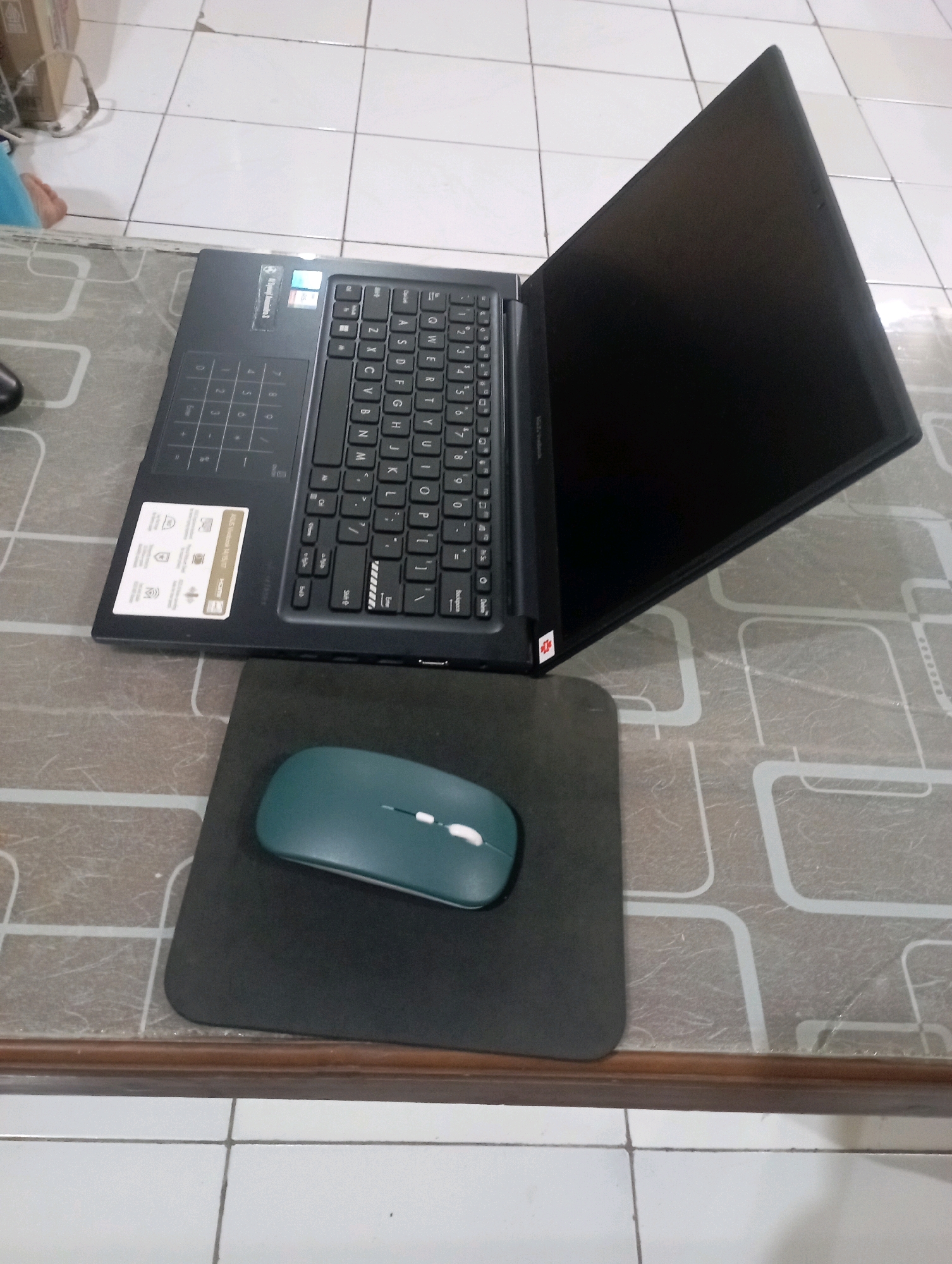 A dark gray laptop is open and angled on a patterned desk, its screen facing upward. To the left of the laptop is a black mousepad with a teal wireless mouse. The desk is positioned on a tiled floor. A box and a person's leg, clad in light blue fabric, are visible in the upper left corner of the image. The overall scene is indoors.