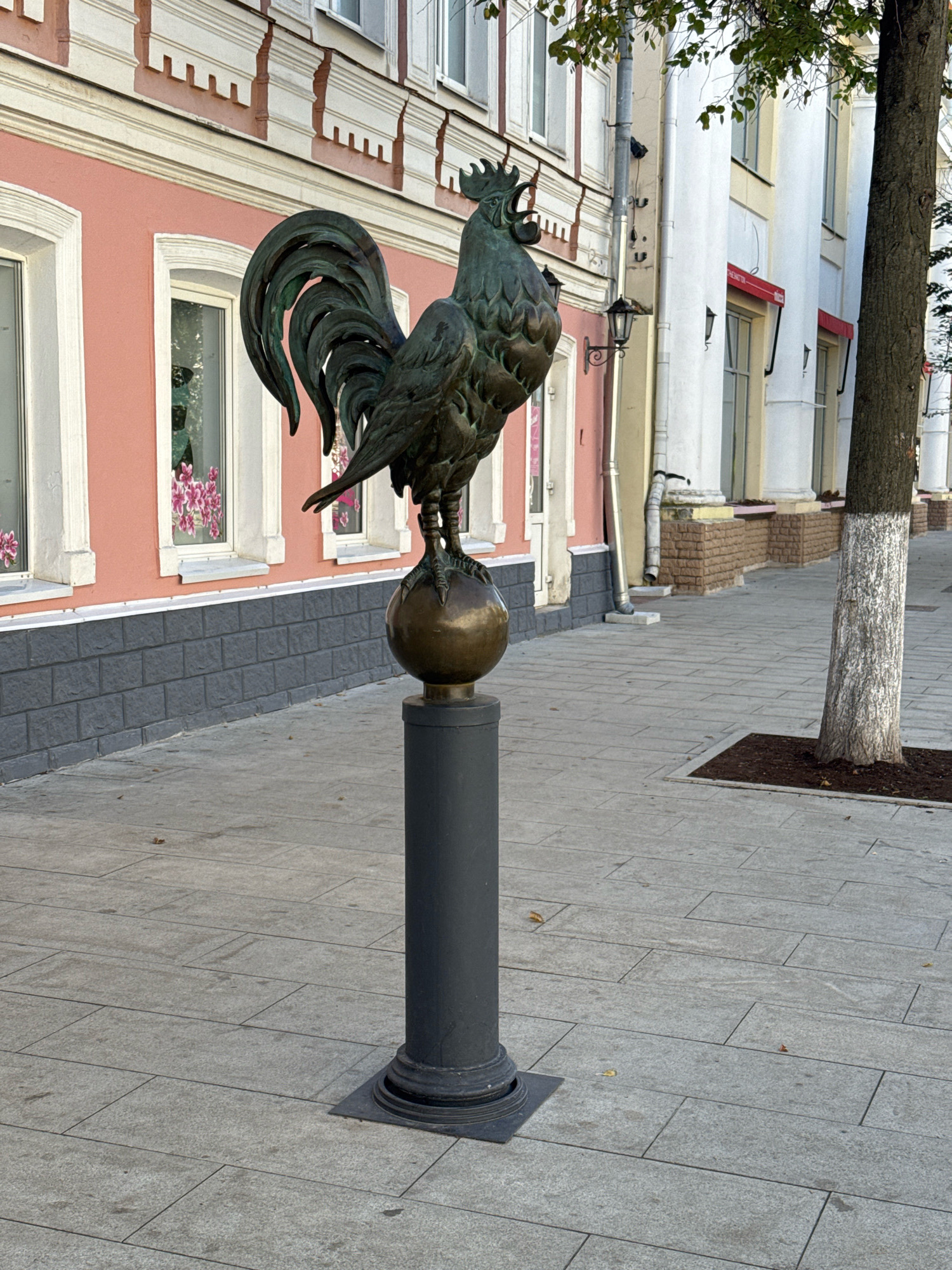 A bronze statue of a rooster stands on a pedestal on a paved sidewalk in front of a building with a pink facade. The building has white-trimmed windows, and the base of the building is clad in dark gray brick. To the right of the statue, a tree with a white-painted trunk grows in a square of dark earth. The sidewalk is made of gray rectangular pavers, and several fallen leaves are scattered across it. The background shows more of the building and its architectural details, including white columns and red awnings over windows. The scene appears to be in a city setting.