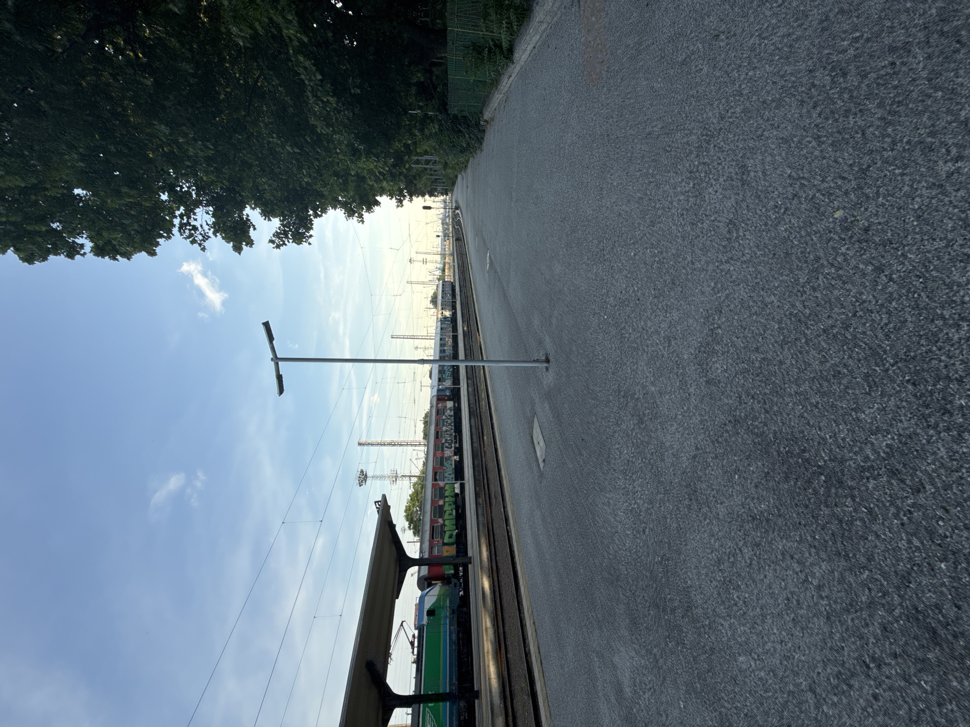 This image depicts a static scene at a train station platform in Burgas, Bulgaria, during daylight hours. The primary view is from a paved platform, likely made of asphalt or compacted gravel, extending towards the background and curving slightly to the right. A tall, dark grey, cylindrical lamppost with a horizontal light fixture stands centrally in the midground of the platform.
To the left of the platform, parallel train tracks are visible, with a multi-car train positioned on the closest track. The front of the train features a locomotive colored primarily green and blue with white accents. Following the locomotive are several passenger cars, predominantly red and white, with extensive, multi-colored graffiti covering their lower sections. Visible graffiti includes the word "CRUMBY" in green block letters on one car. A section of a covered platform structure, supported by dark pillars, partially obstructs the view of the train towards the left.
In the background, beyond the tracks, various railway infrastructure elements are present, including multiple overhead power lines and their support structures, and tall utility poles. To the far right, a dense line of mature trees with dark green foliage runs parallel to the platform, forming a natural boundary. A green mesh fence separates the platform area from these trees. The sky above is a clear, light blue with sparse, wispy white clouds. No individuals are visible in the scene.