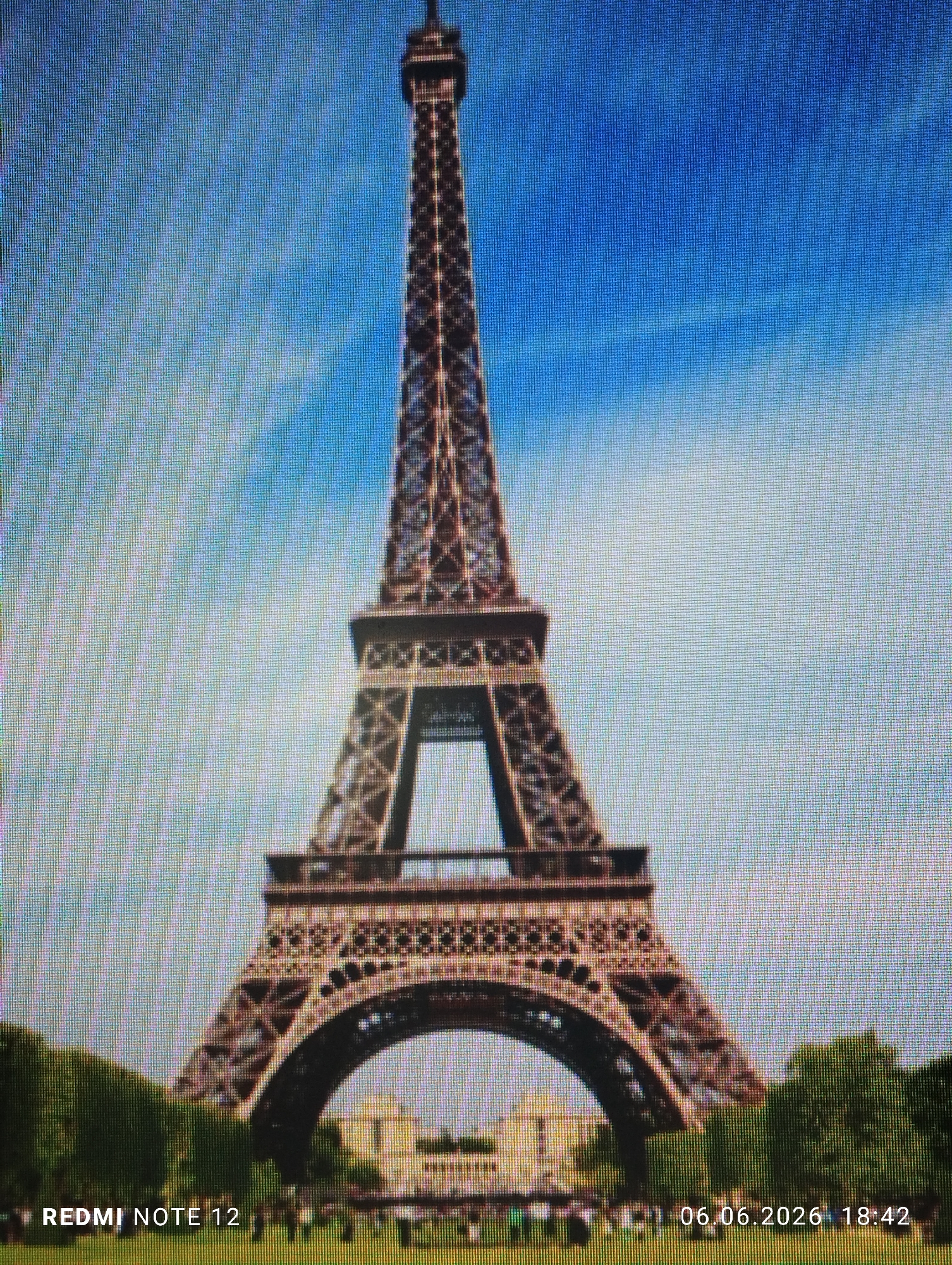 A photograph captures a digital screen displaying the Eiffel Tower, centrally framed against a bright blue sky that transitions to a lighter shade towards the horizon. The iconic iron lattice structure of the tower is detailed, with lush green foliage and distant white buildings visible at its base. The lower foreground depicts a green grassy area populated by numerous indistinct human figures. A distinct pixel grid pattern is visible throughout the image, characteristic of a screen display. Overlaid text reads 'REDMI NOTE 12' at the bottom left and '06.06.2026 18:42' at the bottom right. The location where this image was captured or is being presented is identified as the city of Burgas, Bulgaria.