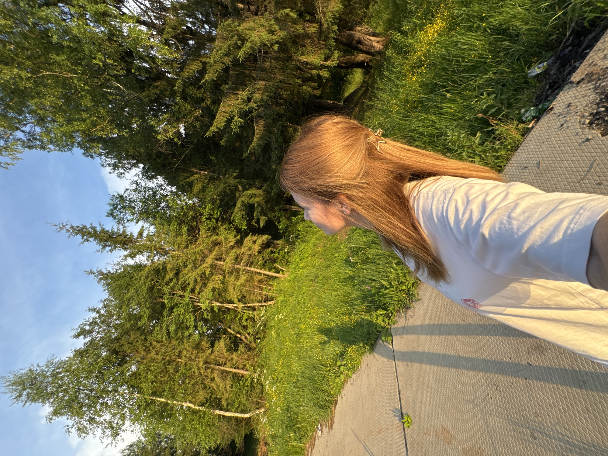 In Вассята, Russia, a young person with long, reddish-blonde hair, adorned with a star-shaped clip, is pictured from the shoulders up, looking upwards. They are wearing a white t-shirt with a faint red logo on the left side. The individual is positioned on a paved surface, possibly a sidewalk or path, with a textured, diamond-like pattern. To the right of the paved area, a vibrant, grassy embankment is covered in lush green vegetation and dotted with small yellow wildflowers. Beyond the embankment, a dense forest of evergreen and deciduous trees stands tall, their branches reaching towards a bright, partly cloudy blue sky. The scene is illuminated by the warm, golden light of a setting sun, casting long shadows from the person and nearby trees onto the pavement and grass.