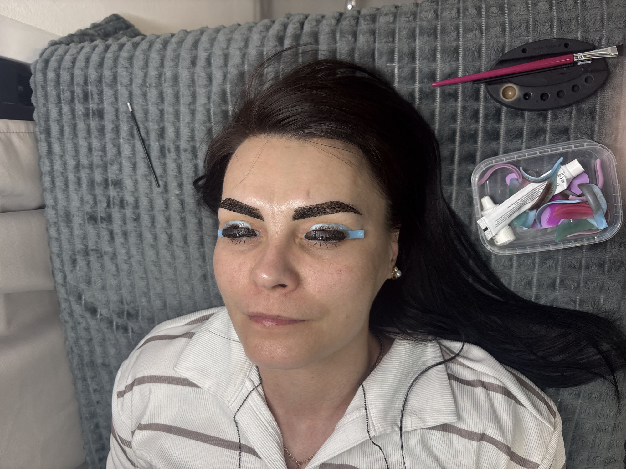 A woman with dark hair is lying on a gray textured surface, possibly a cushion or bed. Her eyebrows appear to be tinted or undergoing a cosmetic procedure. Her eyelashes are being treated with blue lash lift shields that say 'KATYAVINOG' on them.
A container of lash lift products, including tubes and other tools, is visible near her right side. A brush and a small palette are positioned above the container. A makeup brush lies on a black tray.
The woman is wearing a white and gray striped shirt with a collar, and there is a chain necklace visible around her neck. The scene suggests a beauty treatment session, possibly in a salon or private residence. The location is likely Perm, Russia, due to the text on the blue shields that says 'KATYAVINOG', which is a service offered there.