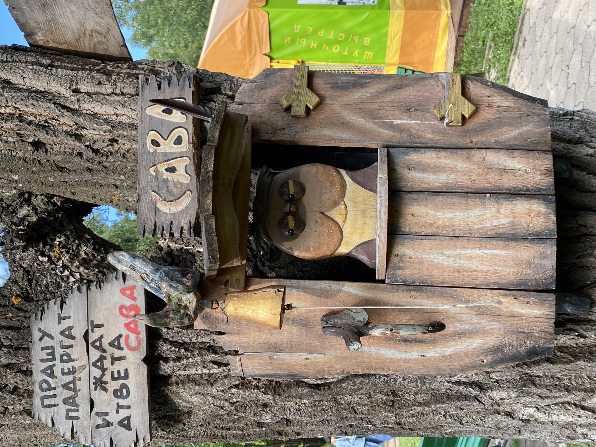 An interactive, whimsical wooden art piece featuring an owl is affixed to a large tree trunk in an outdoor setting, likely a park or recreational area in Chaikovsky, Russia. The central element is a small, rustic wooden house-like structure, from which a carved wooden owl's head with prominent eyes peeks out. Above the owl's opening, a wooden piece is inscribed with "САВА" (SAVA), meaning "owl".
To the left of this owl house, a separate wooden sign is attached to the tree. This sign features text in black and red, reading: "ПРАШУ ПАДЁРГАТ И ЖДАТ ОТВЕТА САВА". This translates to "PLEASE PULL AND WAIT FOR AN ANSWER OWL," indicating the interactive nature of the artwork. A small, golden-colored bell is part of the setup, connected by a string to a wooden handle, inviting visitors to ring it.
The image captures the scene on a bright, clear day, with sunlight illuminating the detailed wooden craftsmanship and the rough texture of the tree bark. In the background, hints of green foliage, a paved path, and a yellow/orange tarp or tent are visible. The overall mood is playful and inviting, encouraging engagement with this unique outdoor installation.