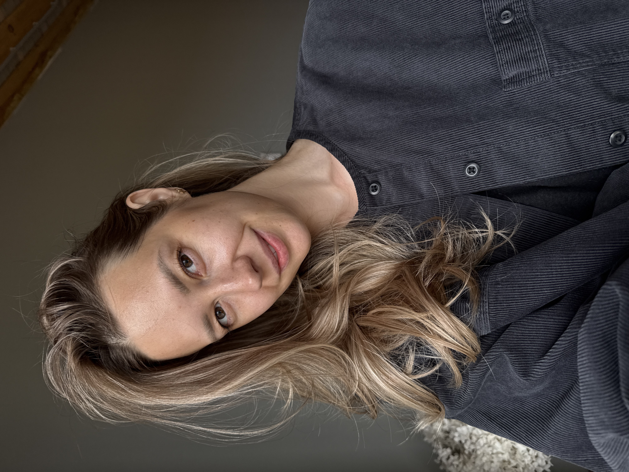 A portrait photograph, taken indoors in Chaikovsky, Russia, features a woman looking directly at the viewer with a soft, gentle smile. Her head is tilted, and her long, wavy hair, a mix of blonde and light brown strands, cascades around her shoulders. She has fair skin and light brown eyes.
The woman is wearing a dark grey or charcoal-colored corduroy shirt or jacket, characterized by its distinct vertical ridges and several visible dark buttons. A small, gold-colored hoop earring can be seen in her right ear.
The background is largely out of focus, dominated by a plain, neutral dark grey wall. In the upper left corner, a partial view of what appears to be a light-colored wooden beam or structural element hints at the interior architecture, possibly a room with exposed ceiling features. A fluffy, light-colored object, perhaps a decorative plant or soft textile, is subtly visible in the lower right corner. The lighting is soft and natural, illuminating her face evenly. There is no visible text in the image.