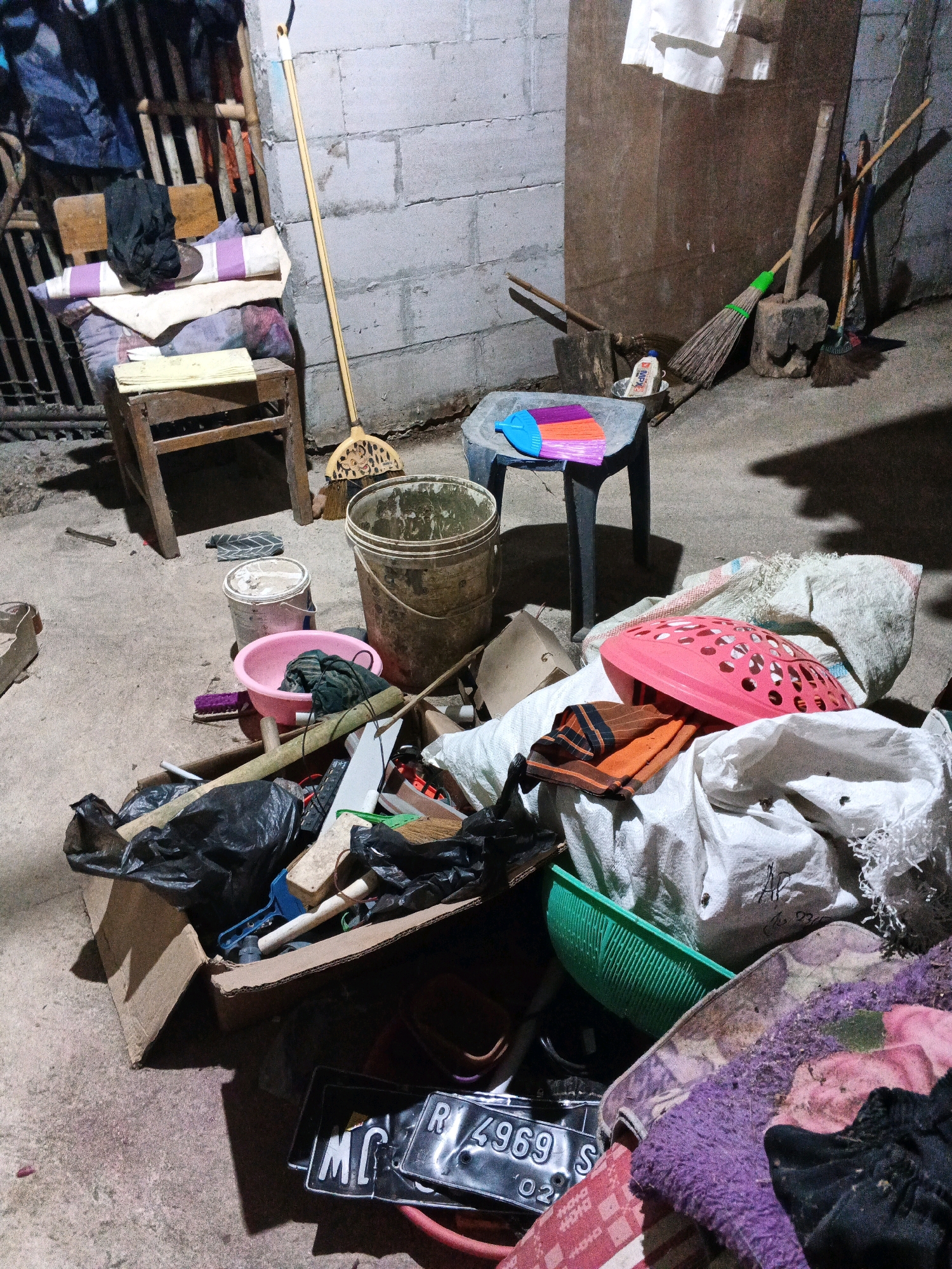 In Sirau, Indonesia, a cluttered storage area is depicted. The scene is dimly lit, with a concrete floor and block-built walls. Piles of objects dominate the foreground, including black trash bags, a cardboard box overflowing with various items such as tools, brushes, and fabrics. Among the debris, a pink laundry basket, a green colander, and several white sacks are visible. To the left, a wooden chair is laden with folded cloths and blankets. Nearby, two buckets, one beige and one pink, sit on the ground. A yellow broom leans against the wall. In the background, several other brooms are propped up against a wooden structure. License plates are scattered on the floor in the lower right corner of the image.