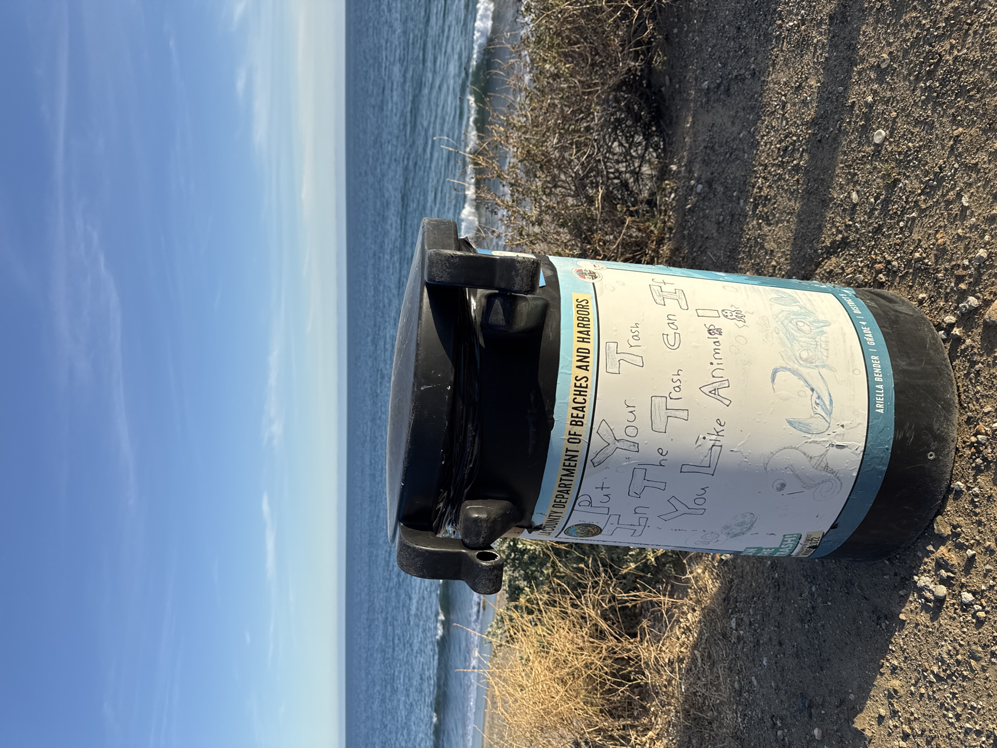 The image shows a black trash can with a white label that reads "Put your trash in the trash can if you like animals!" The trash can is located on a dirt path next to some low-lying vegetation. In the background, there is a view of the ocean and a blue sky. The label includes "COUNTY DEPARTMENT OF BEACHES AND HARBORS" and also "ARIELLA BENDER / GRADE 4 / DISTRICT 3". It is located in Malibu, United States of America.