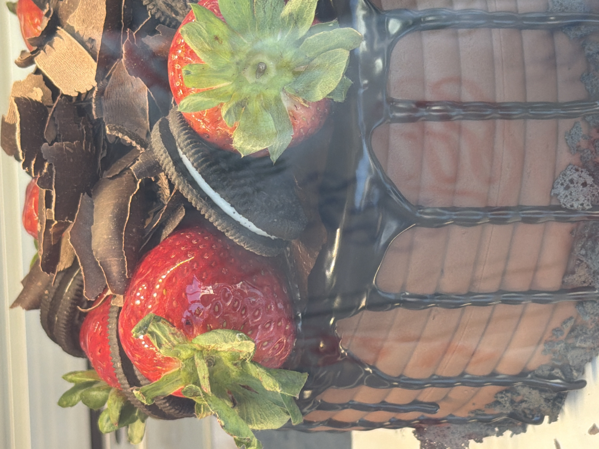 This image captures a vibrant, professionally decorated dessert, likely a cake or large pastry, presented in a vertical orientation. The main subjects are fresh ingredients and confectionery toppings.
The dessert features a rich brown or reddish-brown base, possibly chocolate or red velvet, with a distinct texture on its side, perhaps from a patterned frosting or design. It is elaborately garnished with several bright red strawberries, each retaining its green leafy calyx. Interspersed among the strawberries are numerous dark chocolate shavings, creating a rustic and appealing texture around the base of the fruit. Also visible are several dark chocolate sandwich cookies, resembling Oreos, with their white cream filling peeking out, adding to the dessert's indulgence. A glossy, dark chocolate drizzle or glaze cascades down one side of the cake, forming a grid-like pattern against the underlying surface.
No human subjects or direct activity are visible; the image is a static portrayal of the finished dessert. The lighting appears bright and natural, suggesting it was taken during the daytime, possibly in an indoor setting like a bakery or cafe display case. The background is softly blurred, keeping the focus entirely on the dessert. There is no visible text. While the image itself does not provide specific visual cues for Fethiye, Turkey, this kind of decadent dessert would be a delightful offering in a local patisserie or restaurant in such a scenic location.