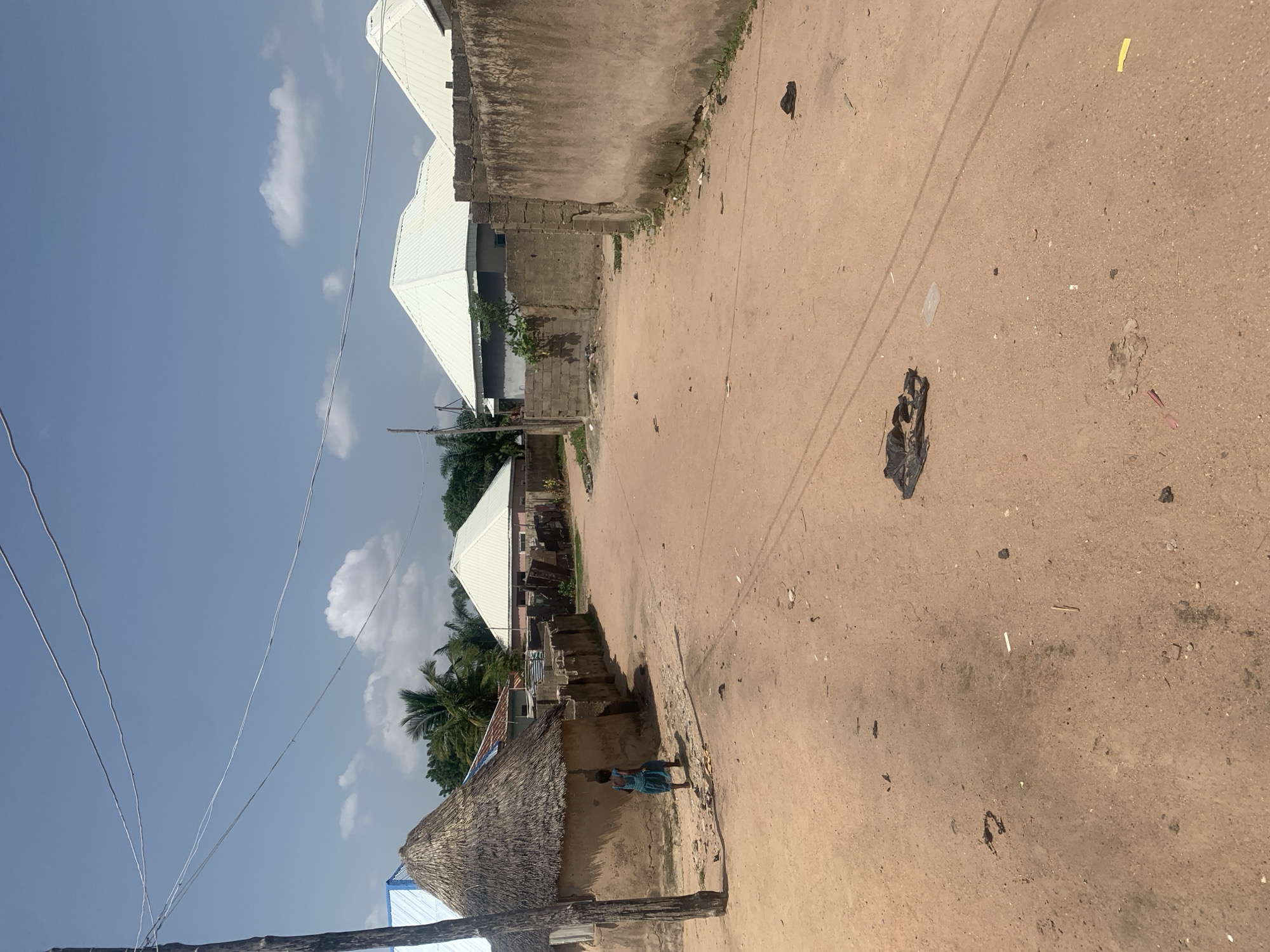 The image depicts a street scene in Jalingo, Nigeria. In the foreground, a dirt road stretches towards the right, with scattered debris including black plastic bags, a piece of yellow plastic, and some grey clumps of dirt. A single power line runs diagonally across the scene. On the left side of the road, a row of buildings with white, angled roofs is visible. One building at the very front has a thatched roof, and a person in a blue and white striped dress stands near its entrance. Palm trees are visible behind the buildings. The sky above is blue with scattered white clouds, suggesting it is daytime with clear weather. The architecture and vegetation suggest a tropical or sub-tropical environment. The scene appears to be a residential or semi-commercial area. There is no visible text in the image.