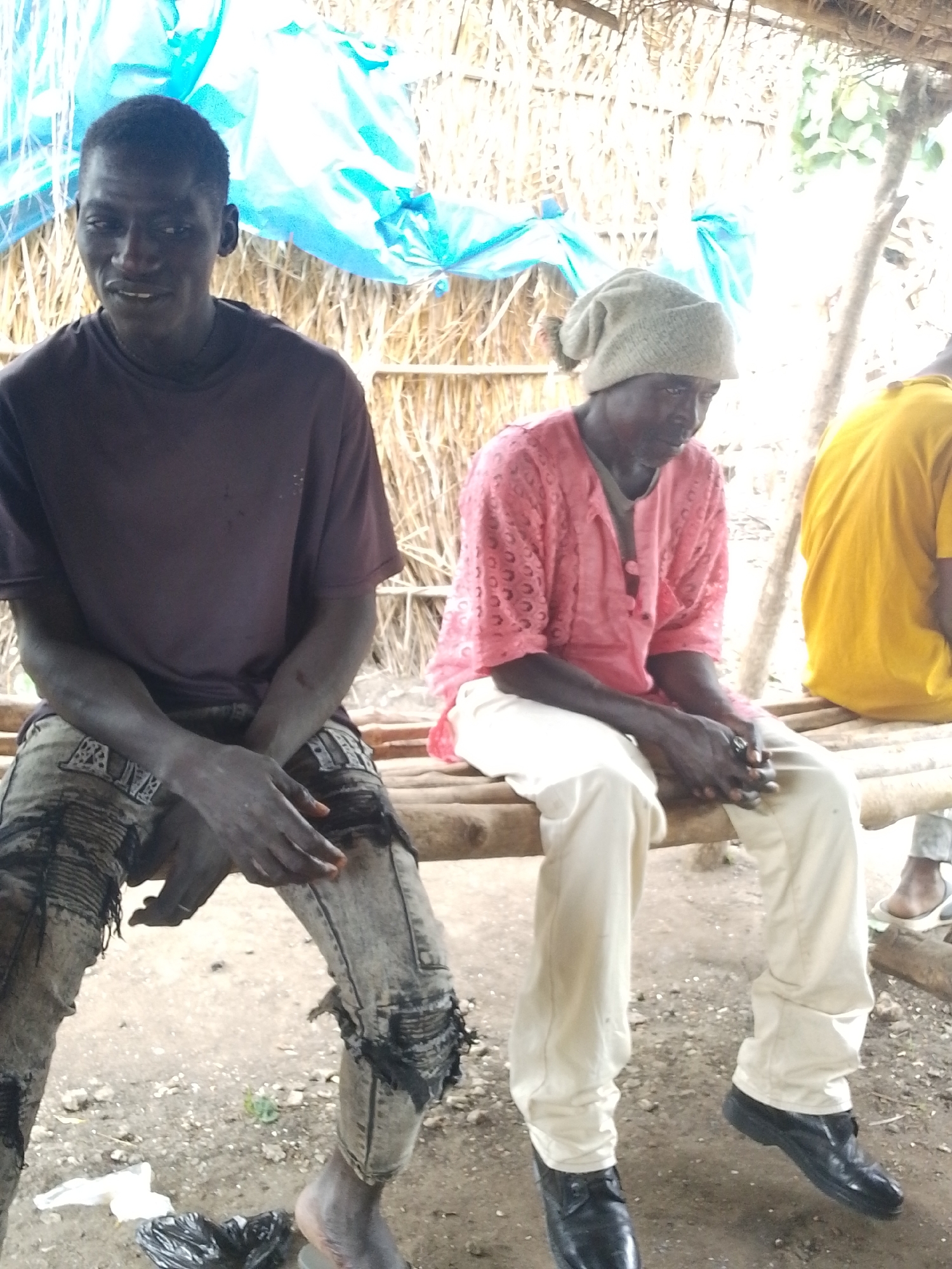 Two men are sitting on a wooden bench outdoors, possibly in Otulu, Nigeria. The man on the left is wearing a dark t-shirt and ripped, distressed jeans. His hands are clasped in his lap. He is looking towards the camera with a neutral expression. The man on the right is wearing a light-colored knit cap, a pink patterned shirt with buttons, and cream-colored pants. His hands are also clasped in front of him. He is looking down with his head slightly bowed. In the background, there is a thatched roof structure with some blue tarp visible. The lighting suggests it is daytime. The ground is covered in dirt and small stones. To the far right, part of another person wearing a yellow shirt is visible, also sitting on the bench. The scene appears to be informal and casual. There is no visible text in the image.