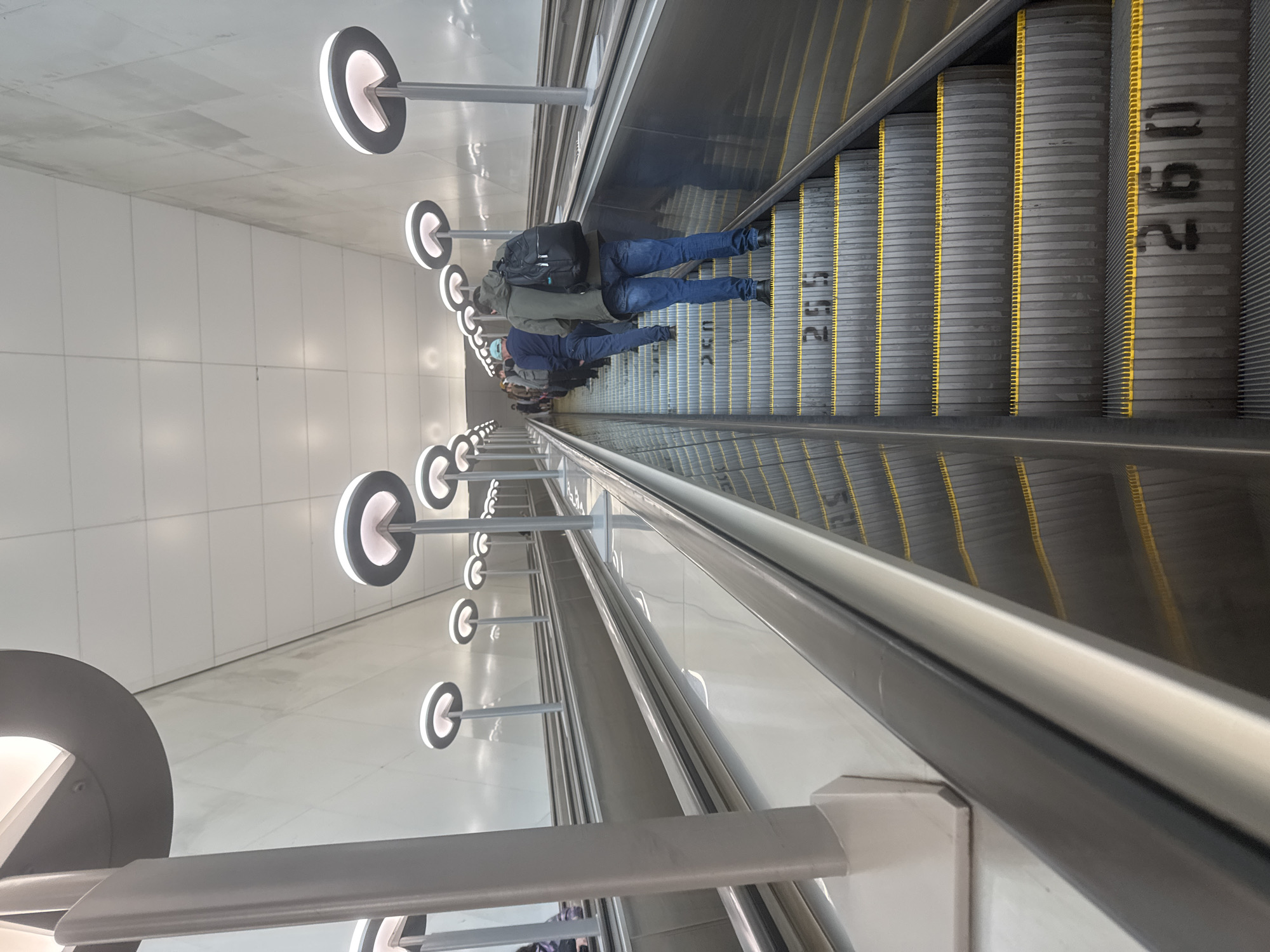 An interior shot shows multiple escalators moving upwards, with several people ascending. The escalators are wide and gray, with yellow safety markings and black numerical indicators on the steps, reading "255" and "260". The walls and ceiling are light-colored and modern, and sleek, circular light fixtures are mounted on tall poles along the escalators. On the rightmost escalator, a person in a green hooded jacket and blue jeans with a black backpack is in the foreground. To their left, another person in a dark blue shirt and light blue cap is visible. The overall scene suggests a public transportation hub or a large, modern building. The escalators appear to be part of a large underground passage in Moscow, Russia.