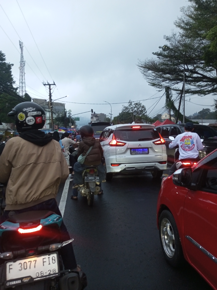 The image depicts a crowded street scene in Dawathang_Dorjibi_ Kashingtsawa, Bhutan, under an overcast sky. The road surface appears damp. Multiple vehicles, primarily motorcycles and cars, are visible in apparent traffic congestion.
In the immediate foreground, facing away from the viewer, is a person on a black Yamaha motorcycle. The rider wears a black helmet adorned with several stickers, including a Turkish flag and text such as "DODTIS," a black hooded garment, and a light brown bomber jacket. The motorcycle's license plate, partially visible, reads "F 3077 FI8" with "06-28" below. To the right of this motorcycle, the right side of a red car is visible, showing a silver multi-spoke wheel and an illuminated brake light.
Further into the scene, directly in front of the foreground motorcycle, a person rides a smaller motorcycle or scooter. This rider wears a dark helmet, a brown puffer jacket, blue jeans, and carries a light blue sling bag. Their license plate reads "F 5850 ZW" with "09-27" below. Immediately to the right of this motorcycle is a white SUV, identified as a Mitsubishi Xpander, with its brake lights illuminated. Its rear license plate reads "F 1157 YS." To the right of the white SUV, another person on a motorcycle is visible from behind, wearing a white long-sleeved shirt with colorful graphics and text, including "BISNIS KECIL KU" and "KOPI & CAFE," and a dark cap.
The left side of the street features various commercial structures or market stalls, with awnings visible. A tall, lattice-structured communication tower stands prominently in the background on the left, alongside a utility pole with numerous overhead wires. On the right side of the street, large green trees line the road, with power lines running through them. A red tent or awning is partially visible in the distant background on the right. Additional vehicles and individuals are visible further down the street, indicating ongoing urban activity.