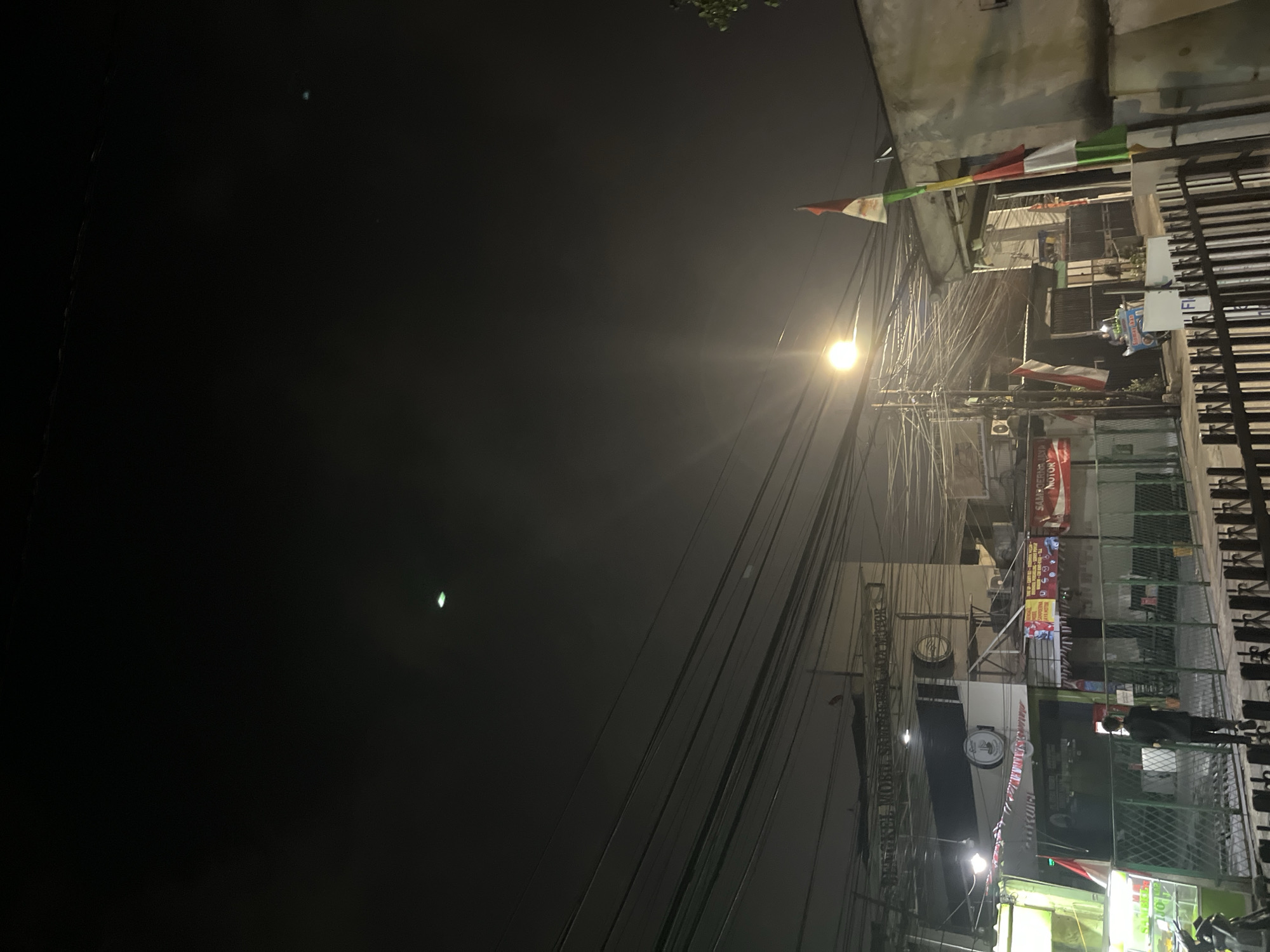 The image captures a nighttime street scene in Central Jakarta, Indonesia, characterized by a dense network of overhead power lines crisscrossing the sky. A street lamp illuminates the area with a warm glow. Several shops and buildings line the street, their facades adorned with various signs, some of which appear to be advertisements. Indonesian flags, red and white, are prominently displayed on poles and buildings. In the foreground, a person dressed in black walks away from the camera, and a metal fence runs along the sidewalk. A small cart, possibly selling food, is parked nearby. The overall impression is of a bustling urban environment after dark, with a distinct local flavor.