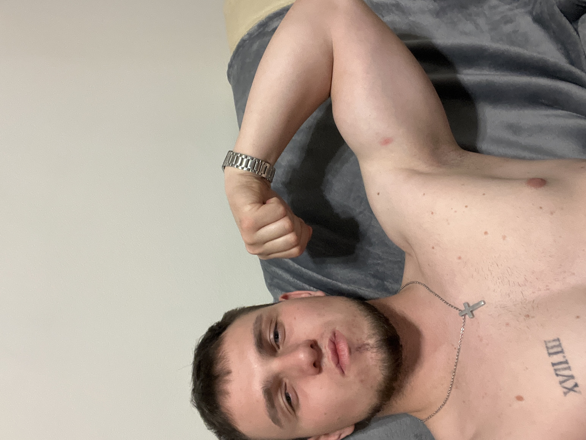 A shirtless man with a beard is lying on a grey blanket, possibly a bed. He has a silver watch on his left wrist and a silver necklace with a cross pendant. A tattoo in Roman numerals is visible on his right chest. The background is a plain white wall. The city and location are Unknown.