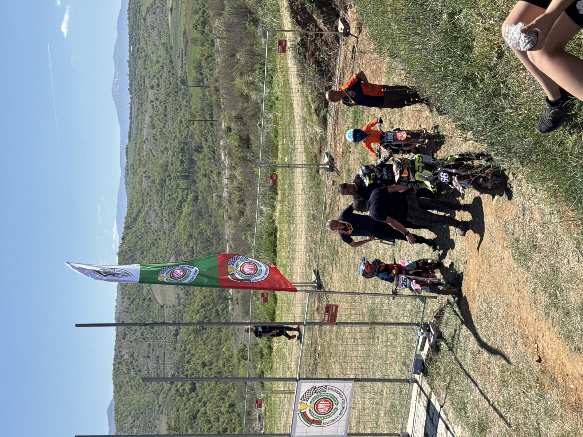 The image captures a motocross event in Selishte, Bulgaria. Several people stand near a dirt track, around three young motocross riders on their bikes, preparing for a race. The riders, dressed in racing gear and helmets, are positioned on their motocross bikes. Adults, possibly coaches or parents, surround the riders, providing support or instruction. A fence separates the track from the spectators and officials. A Bulgarian motocross federation flag is prominently displayed near the track. The background reveals a landscape of rolling hills covered with dense vegetation and a clear blue sky. The scene takes place in daylight, suggesting a race day event.