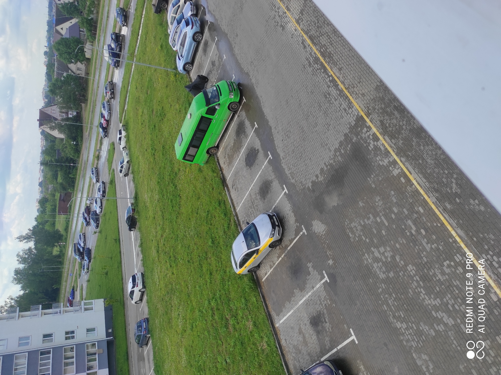 The image shows a parking lot in front of a multi-story building, likely in the city of Maladzyechna, Belarus. The parking lot is paved with grey interlocking bricks, and white lines demarcate parking spaces. Several vehicles are present, including a silver sedan with a yellow stripe, a green minibus, and various other cars parked in the lot and on the adjacent road. The building appears to be an apartment complex with a modern facade. The surrounding environment includes a grassy area, trees, and other buildings in the background, suggesting a residential or mixed-use urban area. The sky is overcast, and there are signs of wetness on the ground, indicating recent rain. The image is captured from an elevated perspective, possibly from a window or balcony.