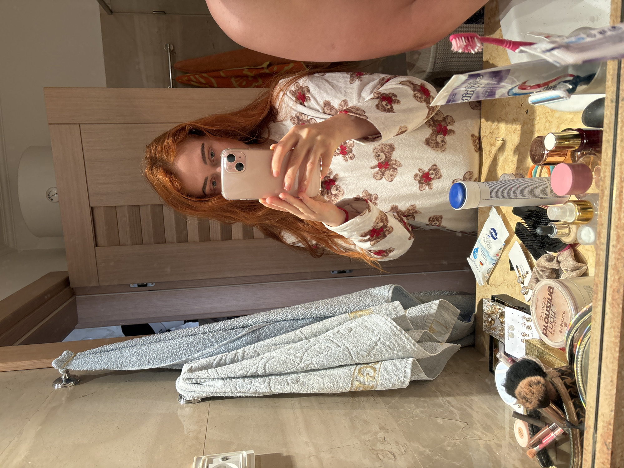 A person is taking a selfie in a bathroom. The bathroom has beige marble floors and walls. In the reflection of the mirror, the person is wearing a white pajama top with teddy bear prints and has red hair. A phone is held up to take the photo. In the background, there is a wooden door and a towel hanging. The sink area is cluttered with makeup and toiletries. The location is a bathroom interior and there is no indication of being in the city of Budva, Montenegro.
