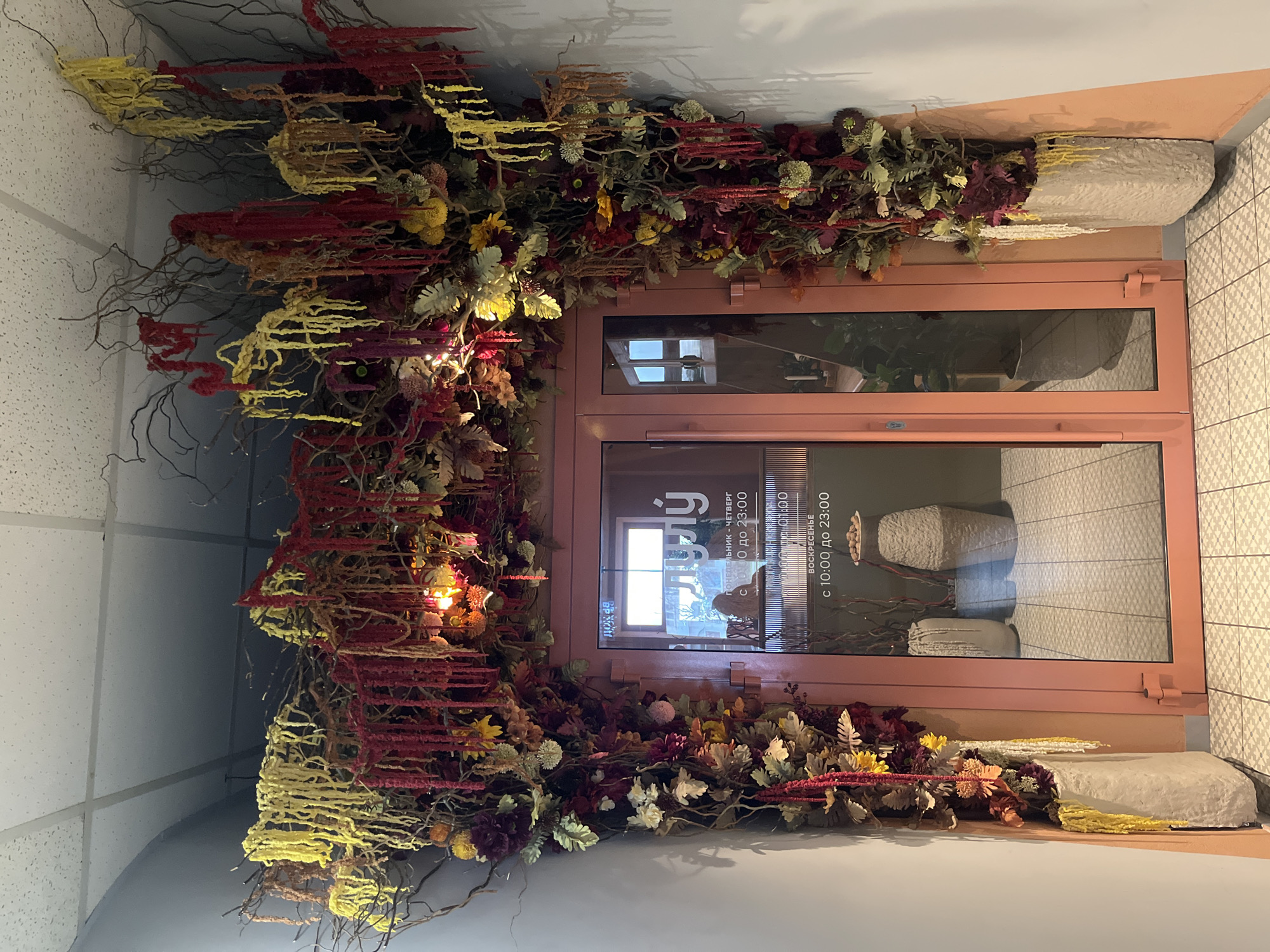 The image shows a highly decorated doorway, likely the entrance to a commercial establishment in Chaikovsky, Russia. The central focus is a double-paneled door with large glass inserts, framed in a muted peach or light pinkish-brown color.
The entire door frame and the surrounding wall space are densely adorned with an elaborate autumnal floral and botanical arrangement. This decoration features a variety of faux dried flowers, leaves, and branches in rich hues of red, yellow, deep purple, and various shades of green and brown. Small, warm string lights are subtly integrated into the foliage, casting a gentle glow. The decorations extend upwards, covering the wall space above the door and along the sides.
Through the glass panels of the door, reflections of the interior are visible, including what appears to be a stylized logo resembling the letter "G" and operating hours. The visible Cyrillic text states: "ПОНЕДЕЛЬНИК - ЧЕТВЕРГ с 10:00 до 23:00" (Monday - Thursday from 10:00 to 23:00) and "ВОСКРЕСЕНЬЕ с 10:00 до 23:00" (Sunday from 10:00 to 23:00).
To the right of the door, a wall with light-colored patterned tiles is visible. Above and below the door (when viewed in correct orientation), a suspended ceiling with square acoustic tiles can be seen. No people are present in the scene, which appears to be indoors. The vibrant yet earthy colors of the decorations suggest a welcoming and aesthetically pleasing entryway. The time of day is not explicitly indicated, but the lighting suggests an indoor setting with ambient or artificial light.
