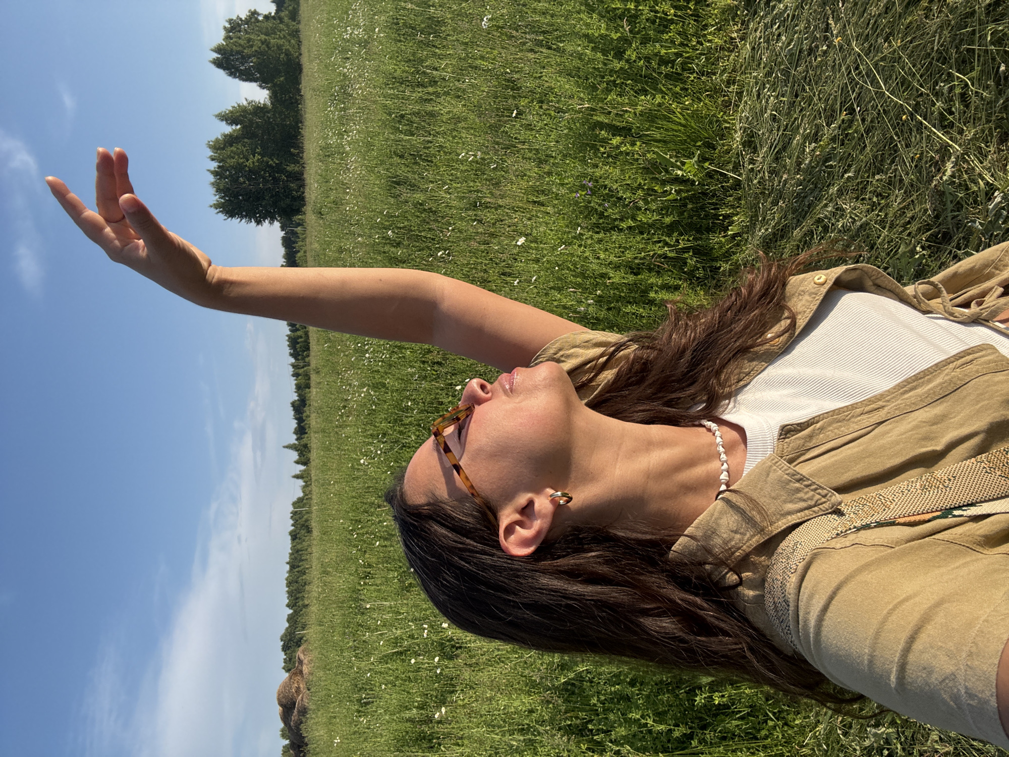 A woman stands in a grassy field with her arm raised towards the sky. The woman is wearing sunglasses, a white top, and a tan jacket. She has long, dark hair and is wearing gold hoop earrings and a beaded necklace. The field is green and appears to have wildflowers scattered throughout. In the background, there are trees and a pile of hay bales. The sky is blue with some clouds. The scene suggests a warm, sunny day. The location is stated to be in the city of Старое Шигалеево, Russia.