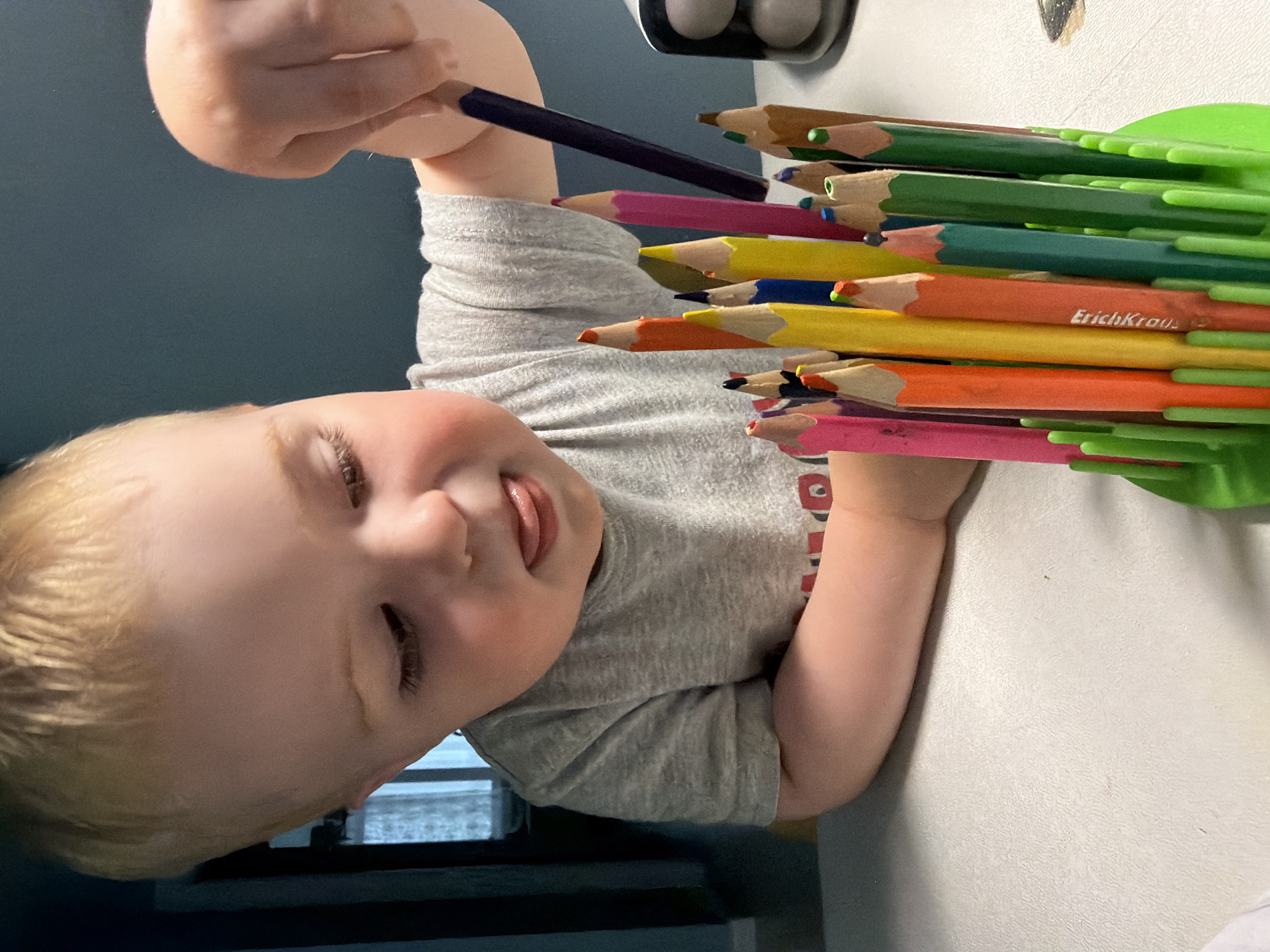A young child with fair skin and blonde hair is holding a blue colored pencil and a container filled with various colored pencils. The child is wearing a gray t-shirt. The container is green and appears to hold the pencils upright. The background includes a blue wall and a white table. The location is impossible to ascertain from the image.