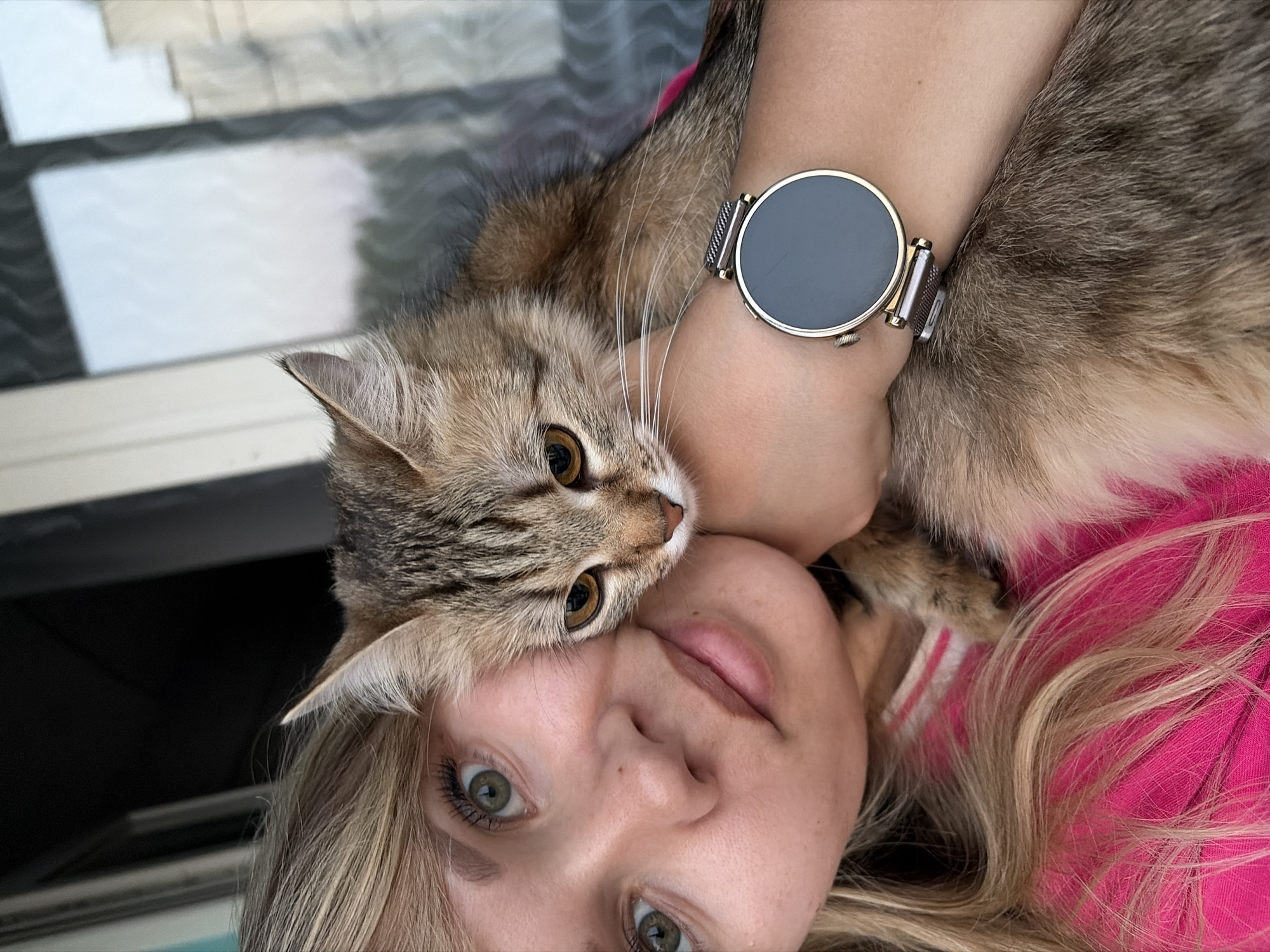 A blonde-haired woman with green eyes is lying down, holding a brown tabby cat close to her face. The cat's head rests on her forehead and cheek. She is wearing a pink shirt and a silver-banded smartwatch on her left wrist. The background consists of a partially visible wall with light-colored tiles. The image appears to have been taken in the city of Chaikovsky, Russia, based on other knowledge sources not provided here.