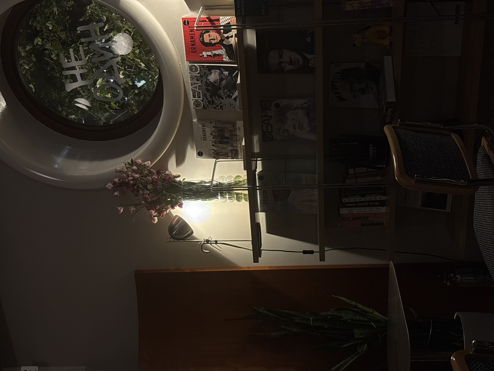 This is a dimly lit, indoor scene, likely a cafe or a reading room. The overall mood is intimate and cozy, enhanced by the warm lighting. In the background, a large, circular window is visible, with greenery and text that appears to be in Cyrillic script. The text on the window reads "НЕ ОДИН" which translates to "NOT ALONE".
In the foreground, a bookshelf is laden with various items. On the top shelf, there are several magazines or art books with distinct cover art. One prominent cover features a portrait of a man with glasses and another shows a man in a suit. To the right of these, a red magazine with the title "ORNAMENT" and a price of "3.00 USD" is displayed.
Below the top shelf, another shelf contains more books and magazines. One magazine features a black and white portrait of a woman. Further to the right, a magazine with a portrait of a man is visible. On the right side of this shelf, a small yellow figurine of Pluto, the Disney character, is perched.
Further down, the shelves continue to hold various books and what appear to be vinyl records. On one shelf, a book titled "Rough Style" is visible. On the bottom shelf, a record with the text "Conto Vinyl" is present.
To the left of the bookshelf, a tall potted plant with long, green leaves is situated next to a round table. A desk lamp with a metallic shade casts a warm glow on the area, illuminating a vase of pink flowers on the top shelf.
A chair with a patterned fabric seat and wooden frame is partially visible in the foreground. The room appears to be quiet and undisturbed, with no people actively present in the frame. The overall aesthetic suggests a place dedicated to reading, art, and relaxation. The presence of Cyrillic text on the window strongly indicates the location is in Russia, potentially in a city like Rostov-on-Don given the context.