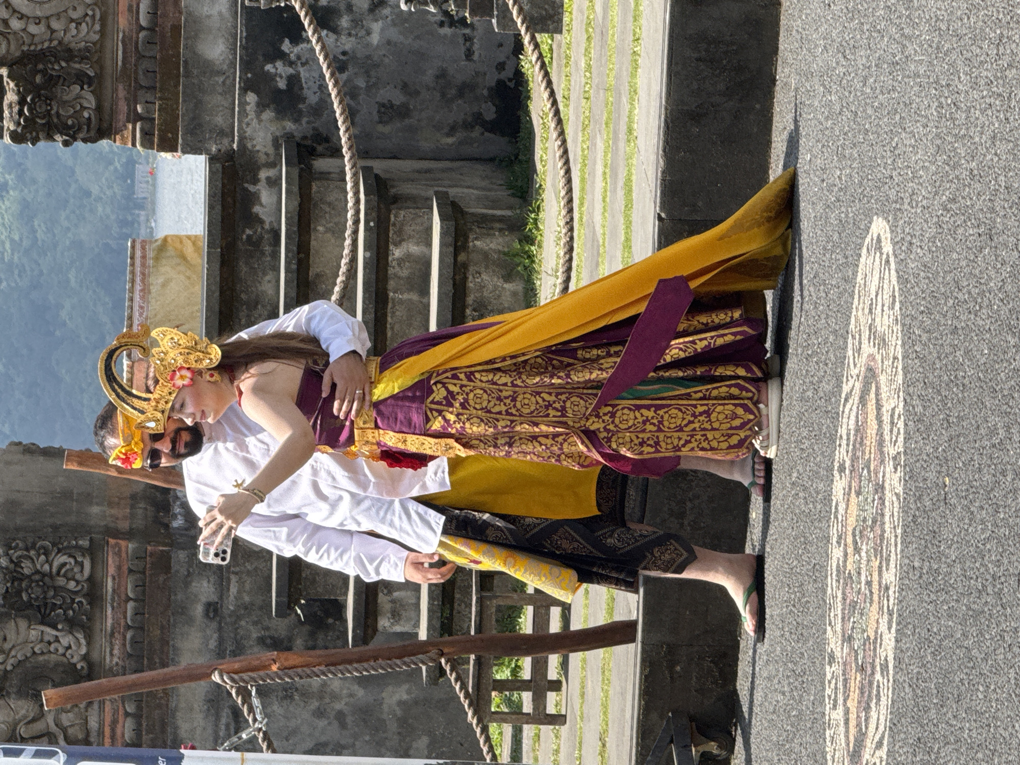 A couple is posing in Baturiti, Indonesia. The woman is wearing a traditional Balinese dress, including a yellow and maroon robe with ornate gold detailing. Her hair is adorned with a gold headdress with a red flower. The man wears a white shirt and traditional black and yellow patterned sarong. He has sunglasses on his face. The woman is holding a cell phone, presumably taking a picture of them. They stand beside gray stone steps and rope railings. The road is made of gray asphalt and painted with a design. Trees and mountains are visible in the background.
