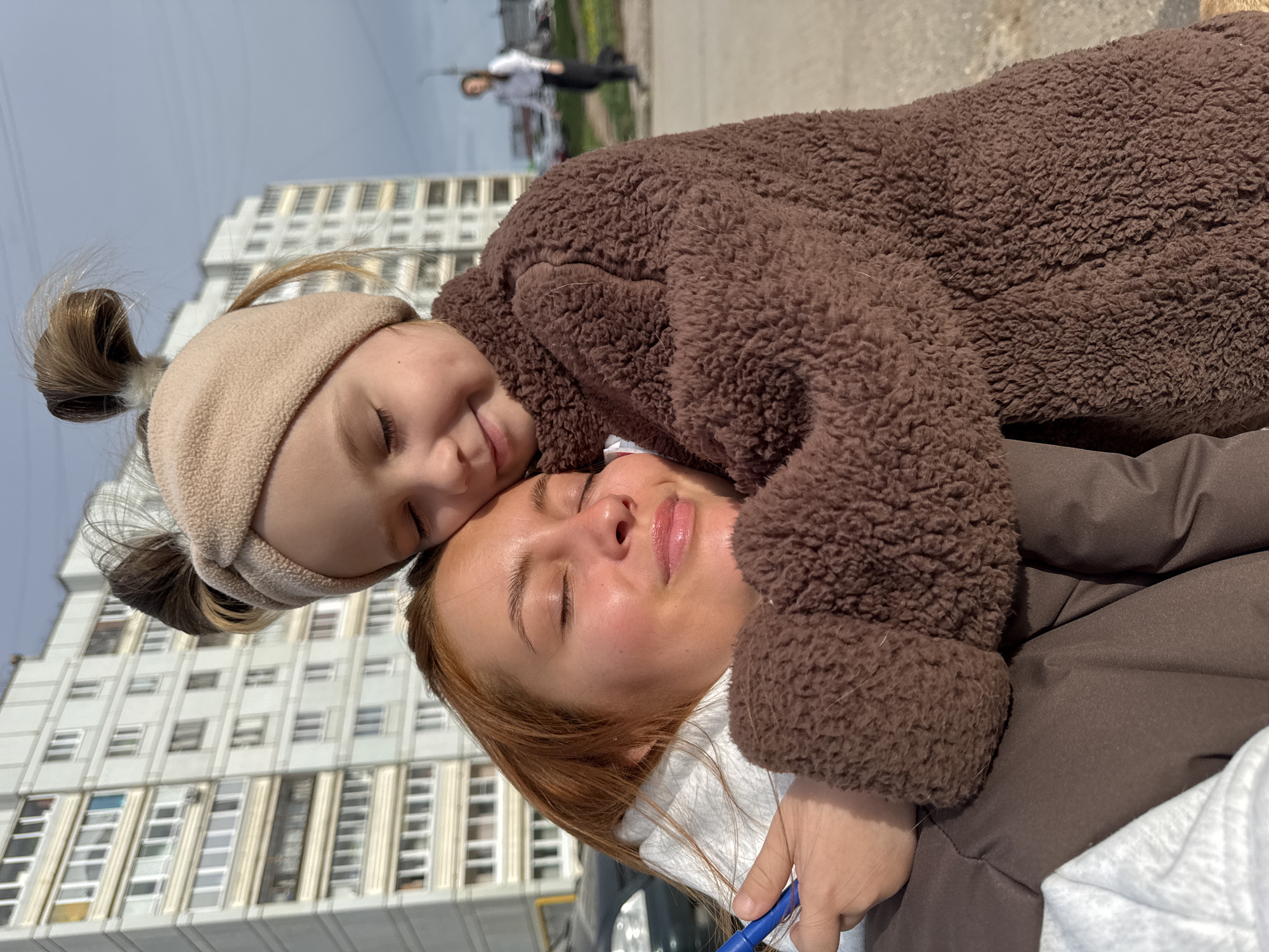 A young girl with a beige headband rests her head on a woman's face in an outdoor setting. The girl wears a brown, textured jacket, while the woman has reddish-brown hair and a white turtleneck visible beneath her jacket. They are positioned in front of a tall, multi-story building with numerous windows, identified as being in Chaikovsky, Russia. In the background, a person is visible walking away from the camera on a paved surface.