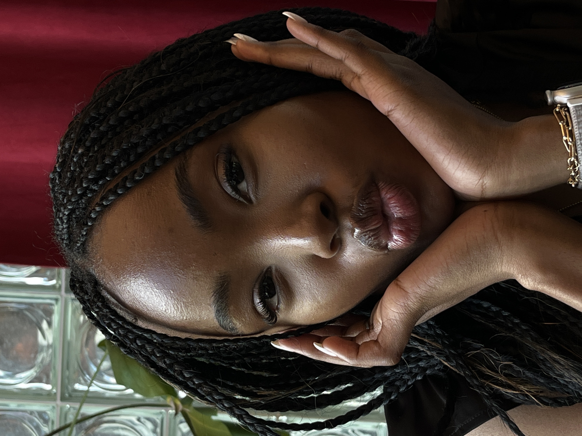 A close-up, eye-level shot shows a young Black woman with braided hair making a kissing face, her hands framing her cheeks, against a dark red backdrop. She is wearing a black top and a gold watch on her left wrist. Her fingernails are painted white. The background includes a blurred view of a plant and frosted glass blocks, suggesting an indoor setting. The lighting is soft and highlights her face. The image is focused on her face and upper body.