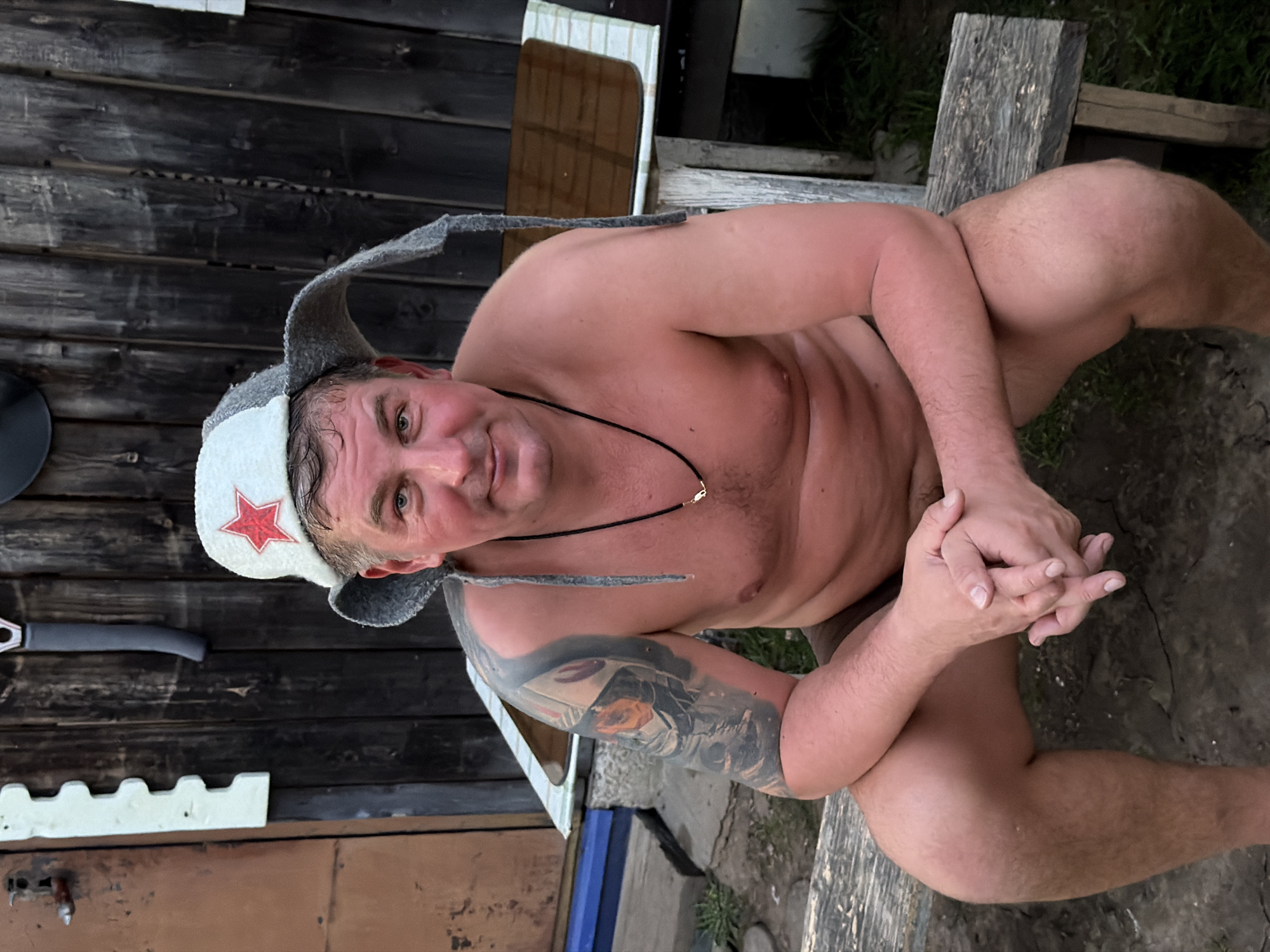 A middle-aged man sits bare-chested on a wooden bench outdoors. He wears a white hat with a red star and grey flaps that extend to the sides. A black necklace adorns his neck, and a tattoo covers his left arm. His hands are clasped together. He looks directly at the camera. The background includes a wooden structure, possibly a building, and overgrown vegetation, indicating a rural or semi-rural setting. This is in Фоки, Russia.