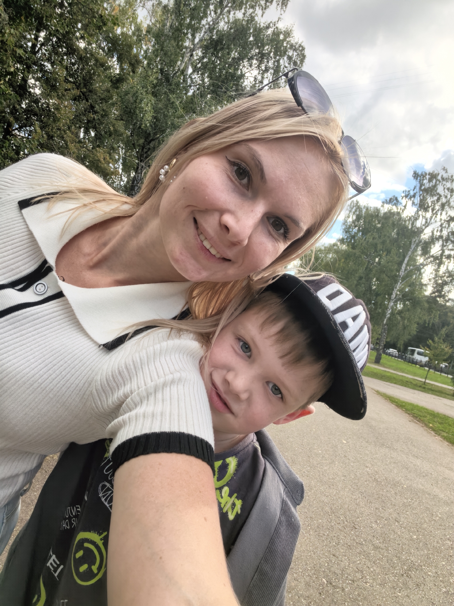 A blonde woman wearing sunglasses on her head and a white sweater with black trim is taking a selfie outdoors. She is holding a young boy in a gray hooded shirt and a black baseball cap with white lettering. The boy is looking at the camera with a slight smile. They appear to be in a park with trees in the background. The road surface is asphalt. No specific details about the city of Sterlitamak, Russia are visible in the image.