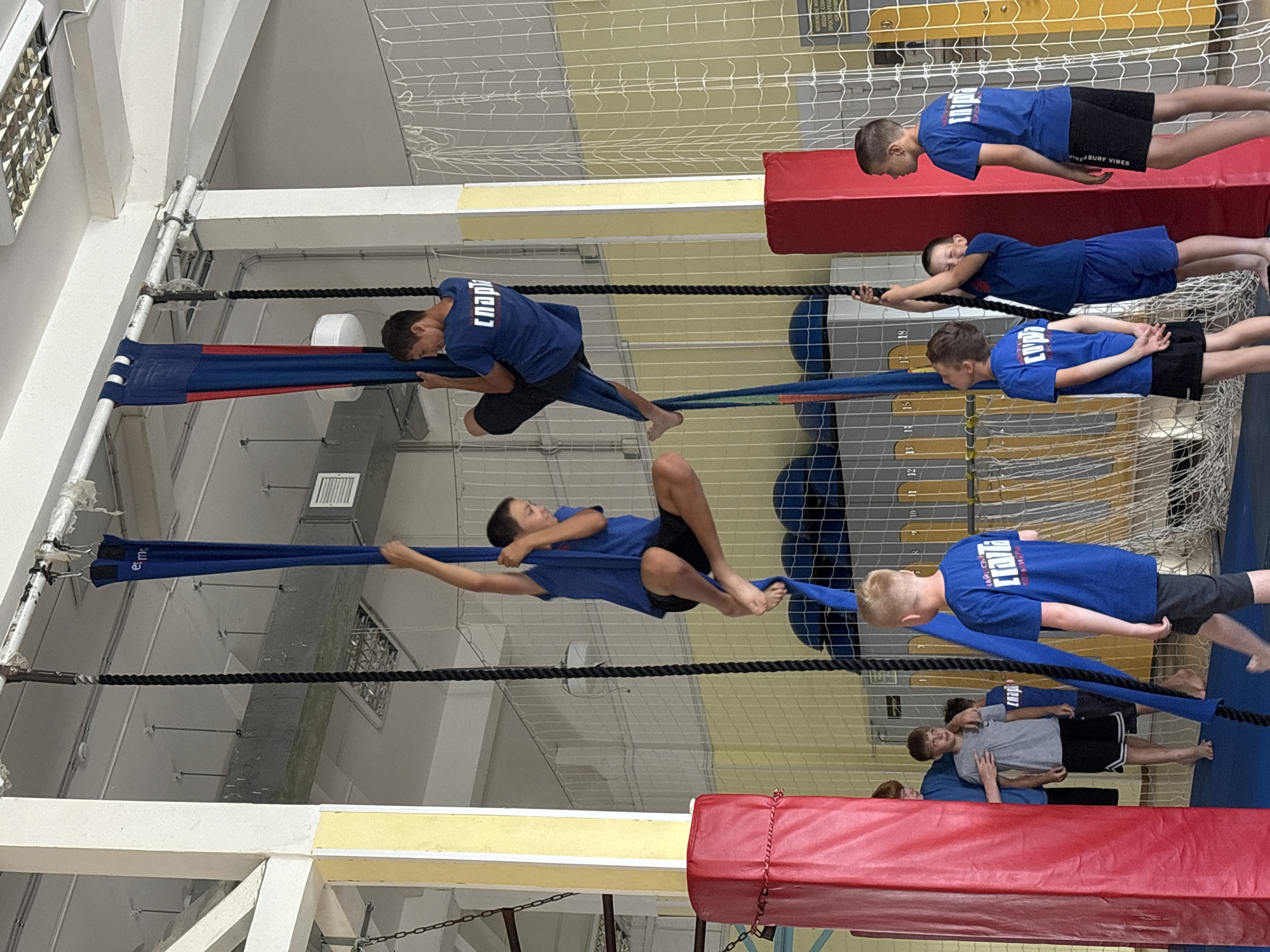 The image depicts a group of young boys engaged in aerial silks and rope climbing activities within a gymnasium. Two boys are suspended mid-air, one on blue aerial silks, and another on a rope. One boy on the silks is in a seated position with his legs crossed, while the other on the rope is also seated. Several other boys are visible on the ground, observing and participating in the activities. One boy on the ground is wearing a blue t-shirt with white lettering that reads "СПОРТ". The gymnasium environment features high ceilings, industrial lighting, and padded mats. The setting appears to be a sports facility, likely a gym or an arena for athletic training. The specific location is a gymnasium in the city of Chaikovsky, Russia, suggested by the Cyrillic lettering on the boy's shirt.