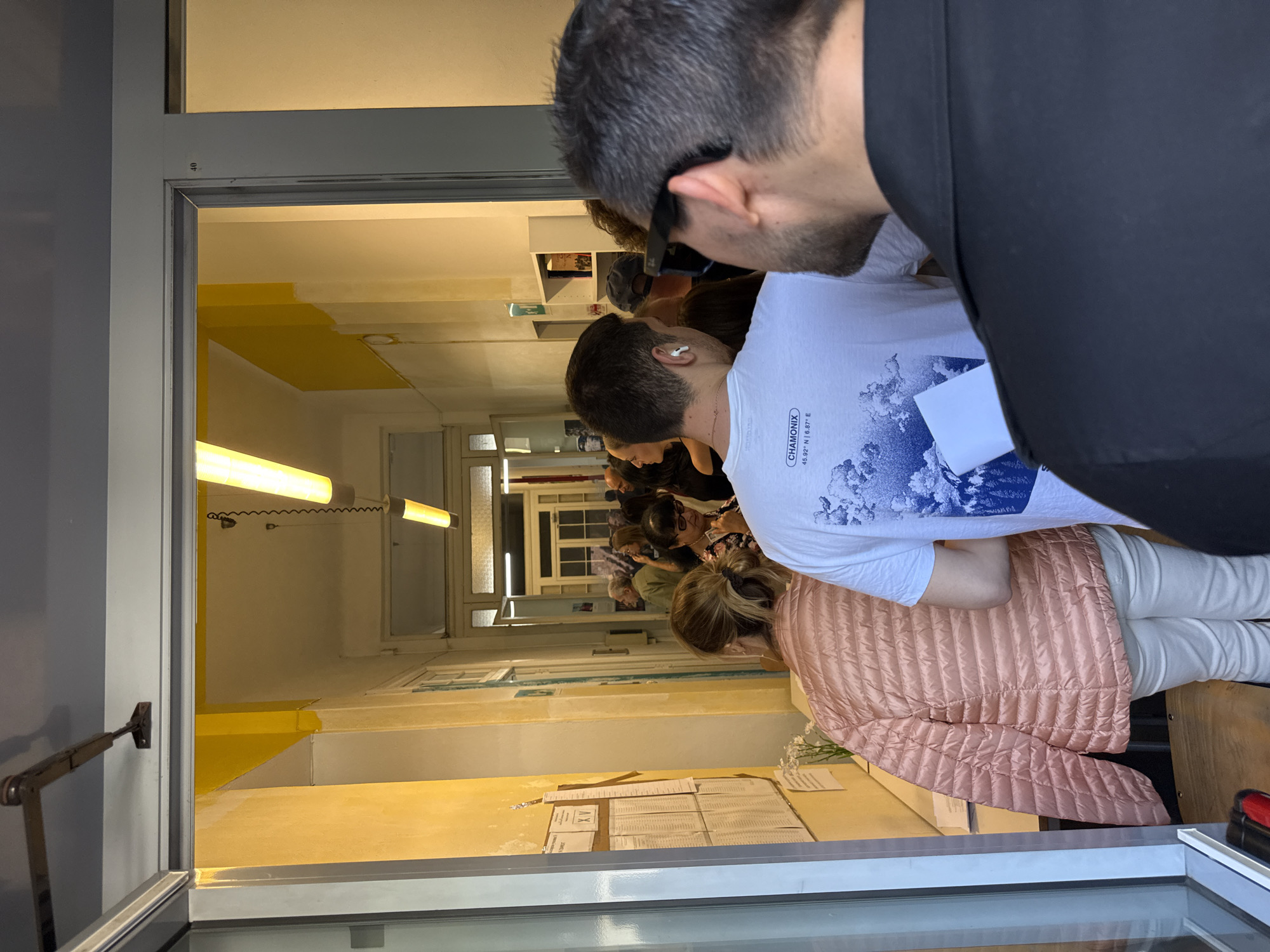 A long, brightly-lit hallway in Milan, Italy is visible through a glass-paneled doorway. People stand in a line, looking at a table with documents posted on the wall. The hallway is painted yellow and white, with long fluorescent lights on the ceiling. Two people are in the foreground, obscuring the view.