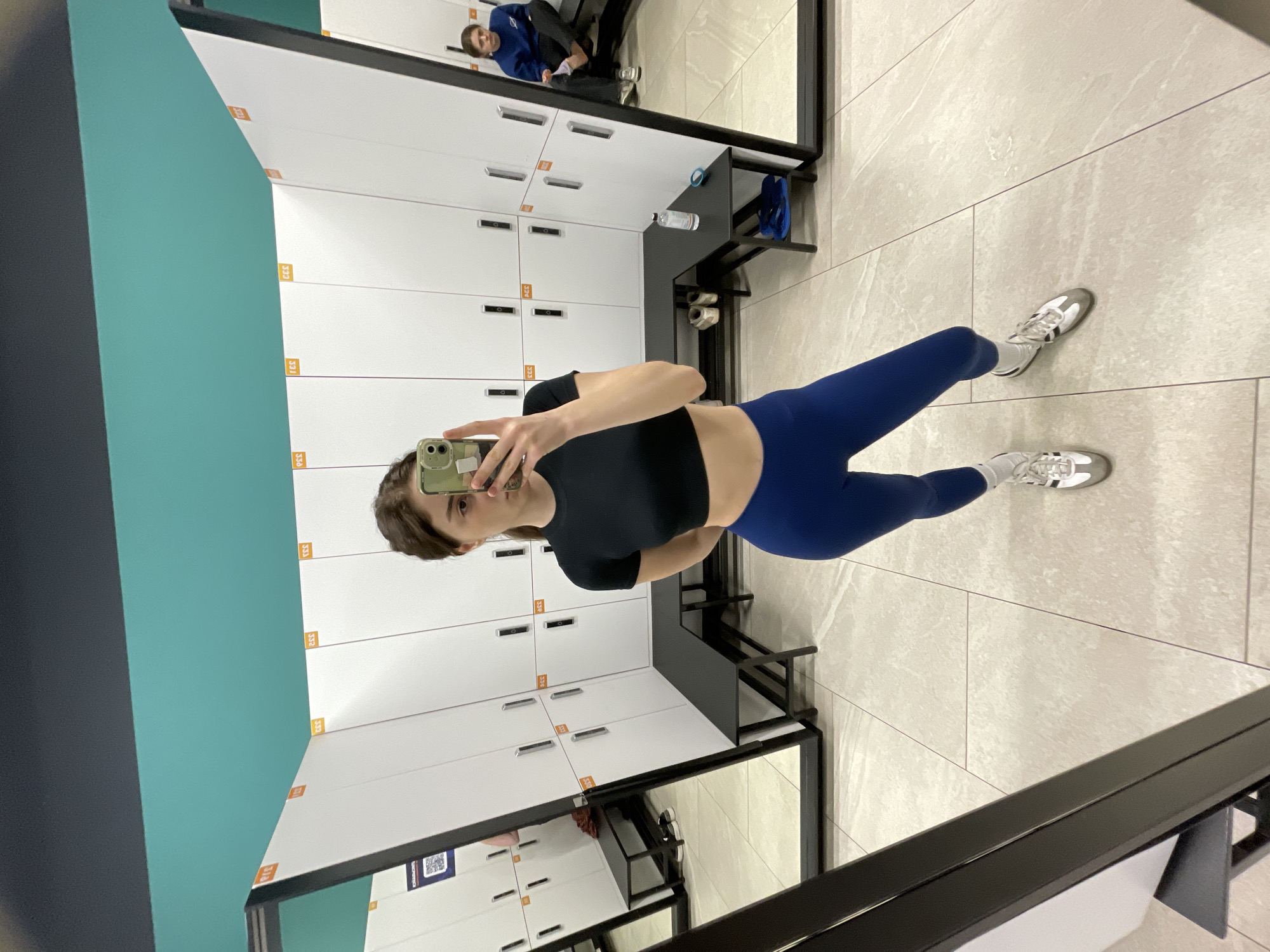 The image appears to be a selfie taken in a locker room. The individual, presumably female, is wearing a black crop top and blue leggings, with white socks and athletic shoes. They are holding a mobile phone to take the picture, with part of their face visible in the phone's display.
The background shows a row of white lockers with orange number labels. There's a bench running along the lockers. The floor is tiled in a light beige color. In the upper part of the mirror reflection, another person is visible, sitting on a bench and putting on shoes.
The image does not provide sufficient information to confirm the location in the city of Moscow, Russia. This information is not discernible from the provided visual data.