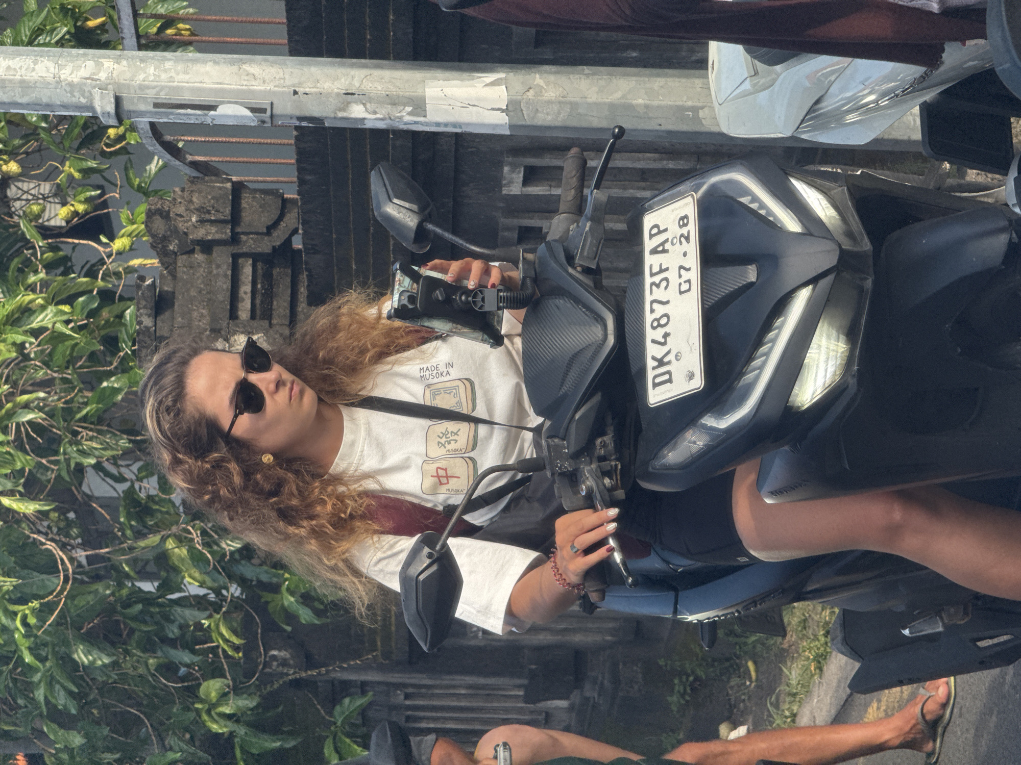 The image depicts a woman with curly hair and sunglasses sitting on a scooter in North Kuta, Indonesia. The scooter has a license plate reading "DK4873F AP 07·28". The woman is wearing a white t-shirt and a red strap across her shoulder. Her hands are on the scooter's handlebars, and she appears to be operating a phone mounted on the handlebars. The background includes foliage and a stone structure. Another person's lower legs and feet wearing flip flops are visible at the bottom of the image.