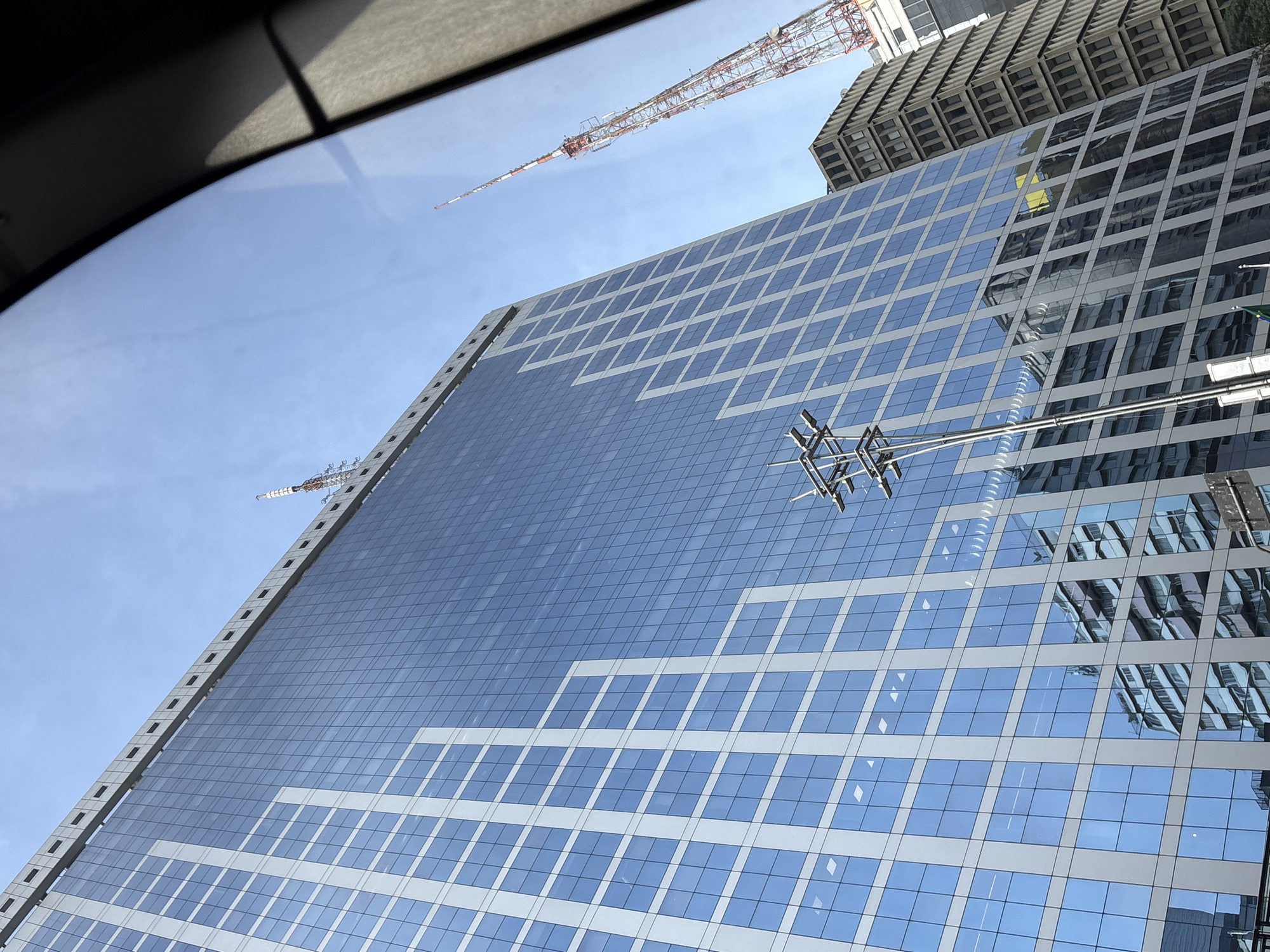 A tall, modern glass-faced skyscraper dominates the frame, its facade reflecting the clear blue sky and other buildings. To the right, a section of a tall, red and white lattice communication tower is visible against the sky. In the background, another older, darker skyscraper with a textured facade stands. The foreground is partially obscured by a dark, curved element, possibly a car window or architectural feature, casting a shadow on the right side of the image. The scene appears to be an urban environment, likely in São Paulo, Brazil, given the presence of such prominent skyscrapers and communication towers. There are no people or visible actions depicted.