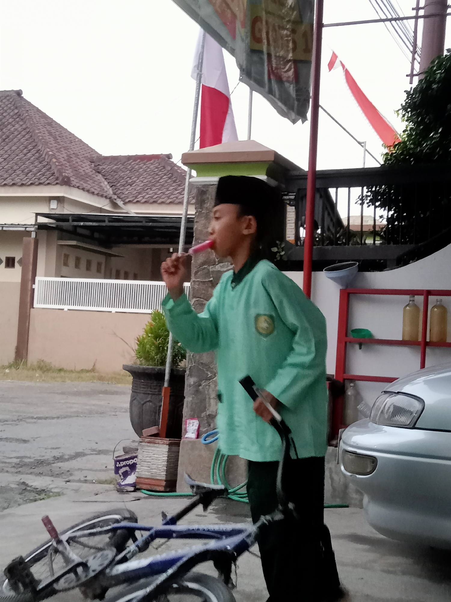 The image depicts an outdoor scene in Simo, Indonesia, during daylight hours. A young boy wearing a black peci (cap) and a light green long-sleeved shirt is standing, holding a pink popsicle to his mouth. He appears to be looking towards the left of the frame. In the foreground, a bicycle lies on its side, with its front wheel turned towards the right. Behind the boy, a silver car is parked to the right, with its headlights visible. Further back, a house with a tiled roof and white fencing is visible. Indonesian flags are displayed prominently in the background. The environment includes paved ground and some vegetation.