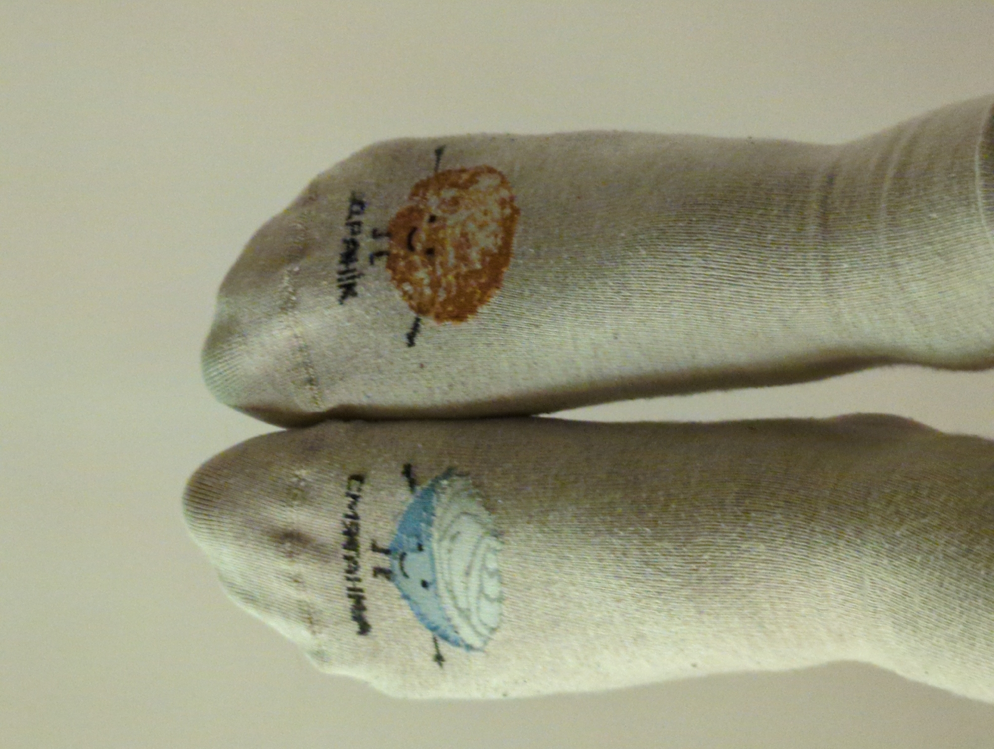 Two white socks are placed side by side on a flat surface. Each sock has a cartoon drawing near the toe area. The top sock features a brown, round drawing with facial features and the word "Apelsin" written vertically next to it. The bottom sock has a blue, round drawing with facial features and the word "Chernika" written vertically next to it. No specific location in Minsk, Belarus, is discernible from the image.