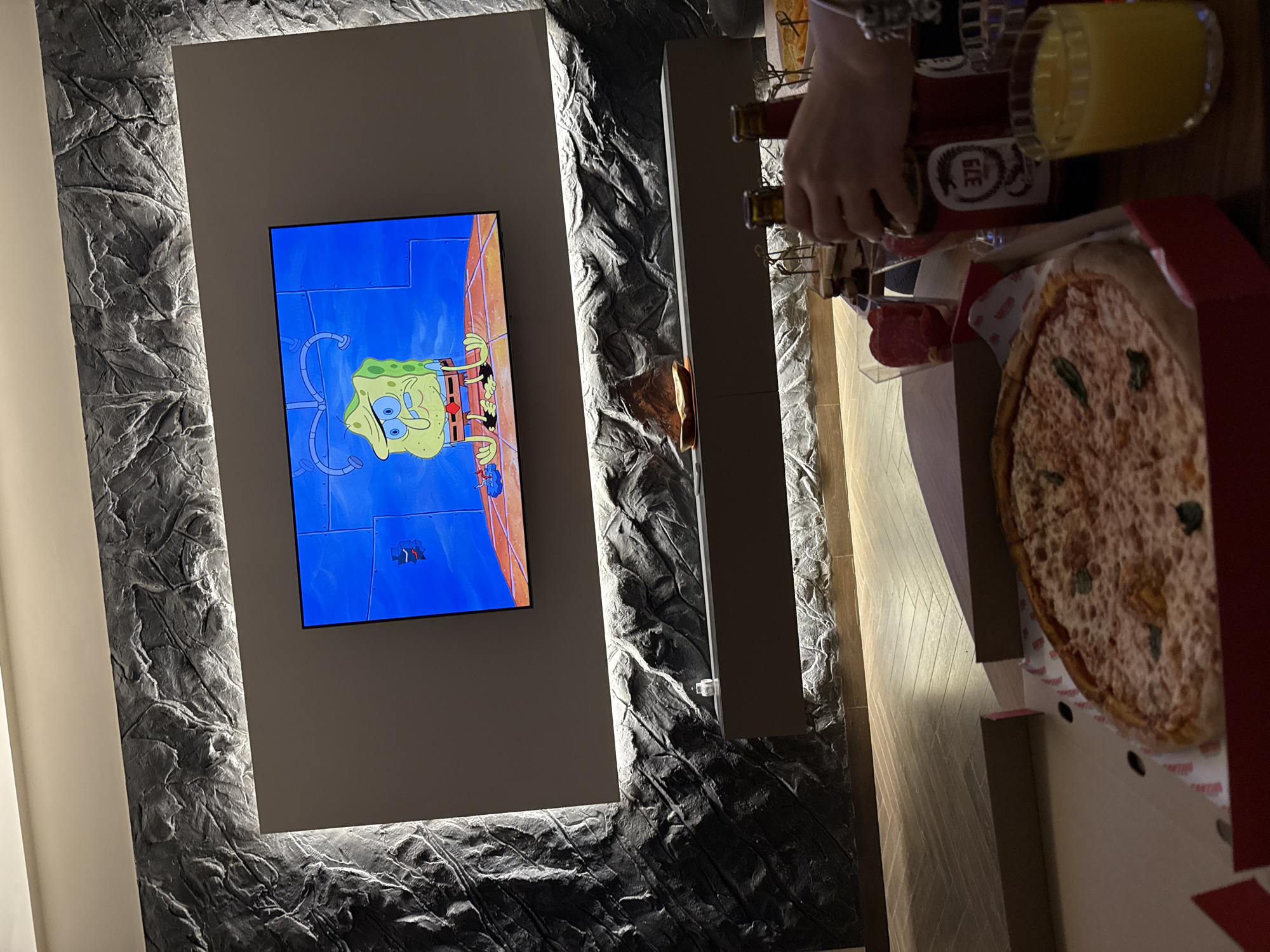 The image, rotated counter-clockwise, captures a cozy indoor setting, likely in Yerevan, Armenia, during the evening. A large flat-screen television is mounted on a striking dark gray wall with a textured, rock-like finish, featuring integrated strip lighting. The TV screen displays an episode of Spongebob Squarepants, with the main character prominently visible.
To the right of the television, a person's hands are visible on a table, holding a red bottle and seemingly pouring its contents. The bottle has "ELLE" printed on its label. Next to it, a glass filled with an orange liquid, possibly juice, is present. A large pizza, still in its red delivery box, takes up a significant portion of the table, and small dishes containing slices of cured meat, possibly salami, are also visible. The overall scene suggests a relaxed evening of dining and entertainment.