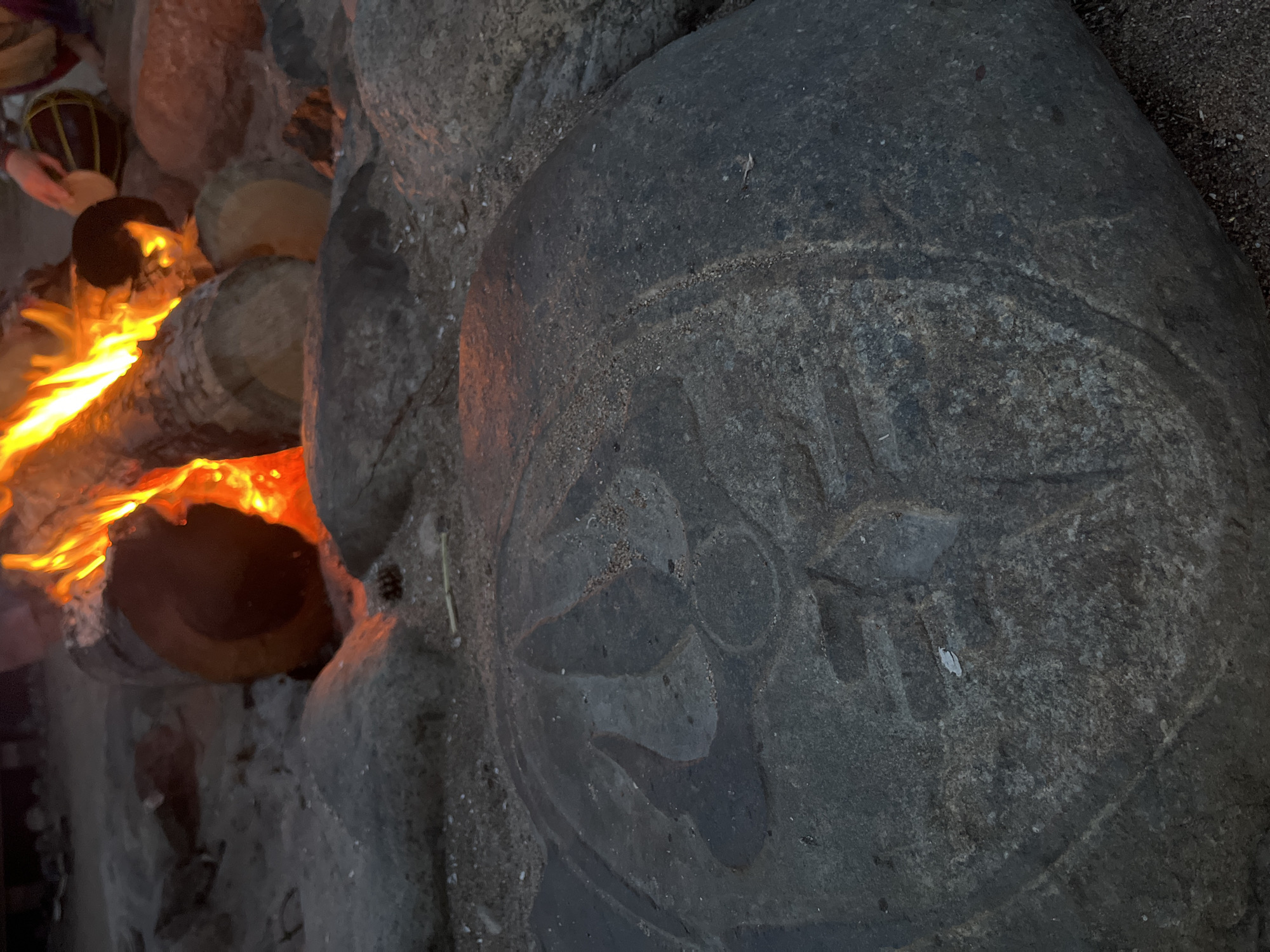 A close-up, low-angle shot focuses on a carved stone in the foreground, its intricate design partially obscured by shadow and sand. The stone appears weathered and sits on a sandy, rocky surface. In the background, a campfire blazes, casting a warm orange glow on several logs and the surrounding rocks. The environment suggests an outdoor setting, possibly a campsite, with a hint of dusk or evening. Two individuals are partially visible near the fire; one is playing a drum.