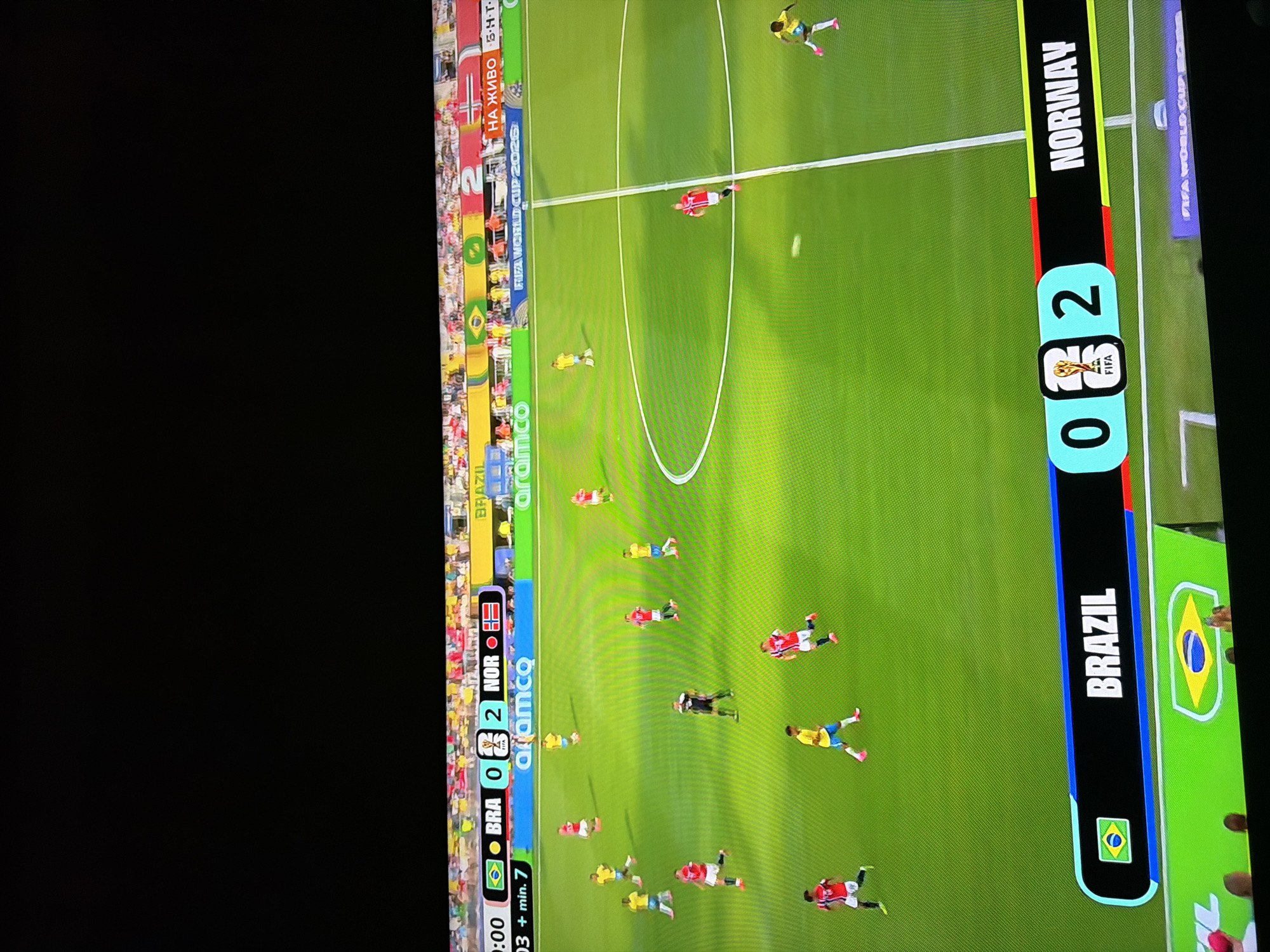 This is a photograph of a soccer match being shown on a television screen. The scoreboard at the top of the screen indicates that Brazil is playing Norway, with Brazil having 0 goals and Norway having 2 goals. The time on the clock is 0:03 + min. 7. The screen displays the FIFA World Cup 2026 logo. The scene appears to be taken from a room where the television is being watched, with a black ceiling visible above the screen. The bottom of the image shows part of a green banner with the Brazilian flag and the words "BRAZIL" and "NORWAY" on either side of the score. There are also spectators visible in the lower part of the image. The photograph does not contain any information about the city of Ognianovo, Bulgaria.