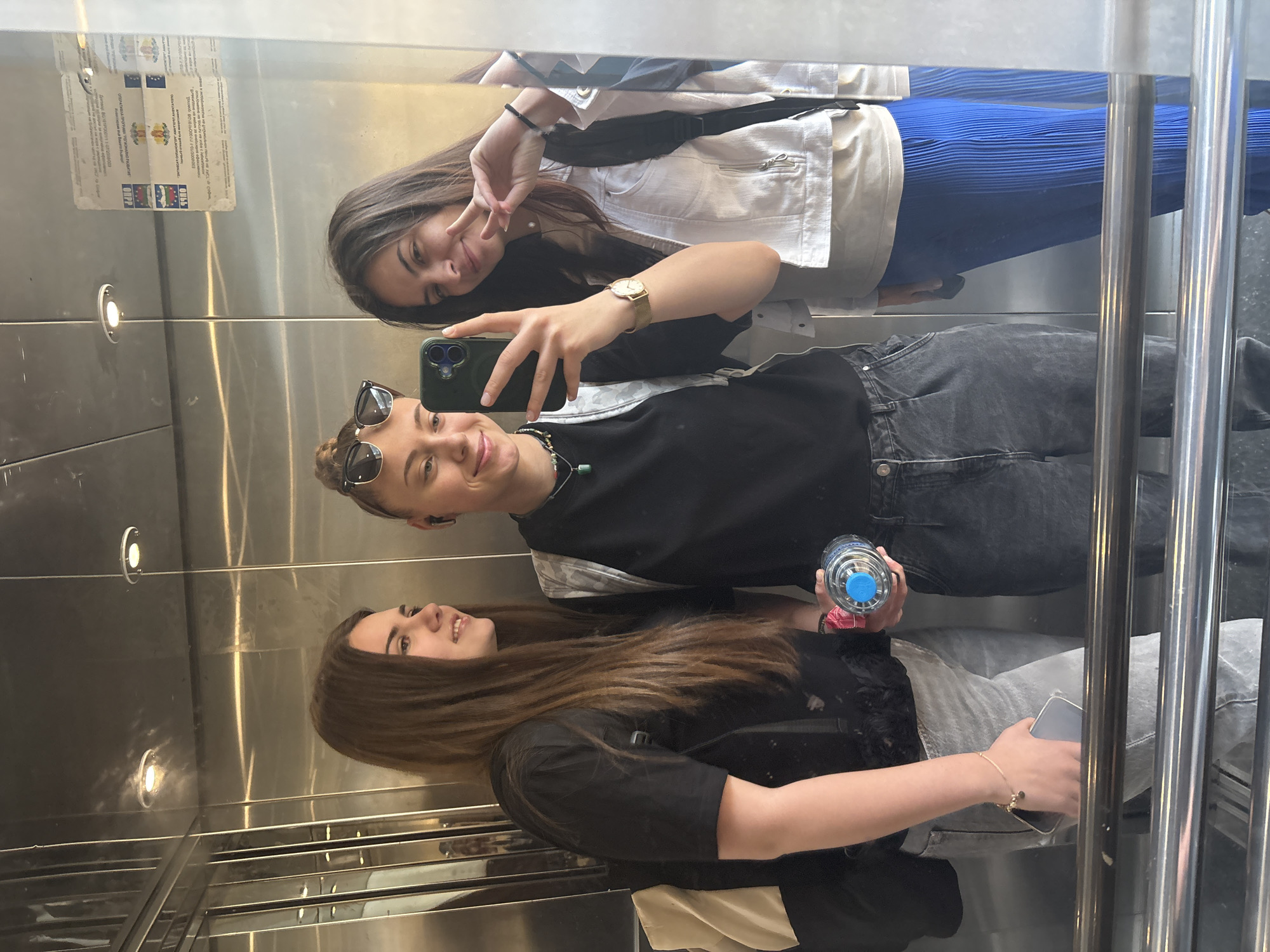 Three young women are taking a selfie in an elevator with mirrored walls. The person holding the phone is wearing sunglasses, a black t-shirt, and grey jeans. One woman is wearing a white jacket, blue pants, and has her hand up in a peace sign. The third woman is wearing a black shirt and grey jeans and holding a cell phone. One of the women is also holding a water bottle. A sticker is visible in the mirror near the top. The elevator is located in Sofia, Bulgaria.