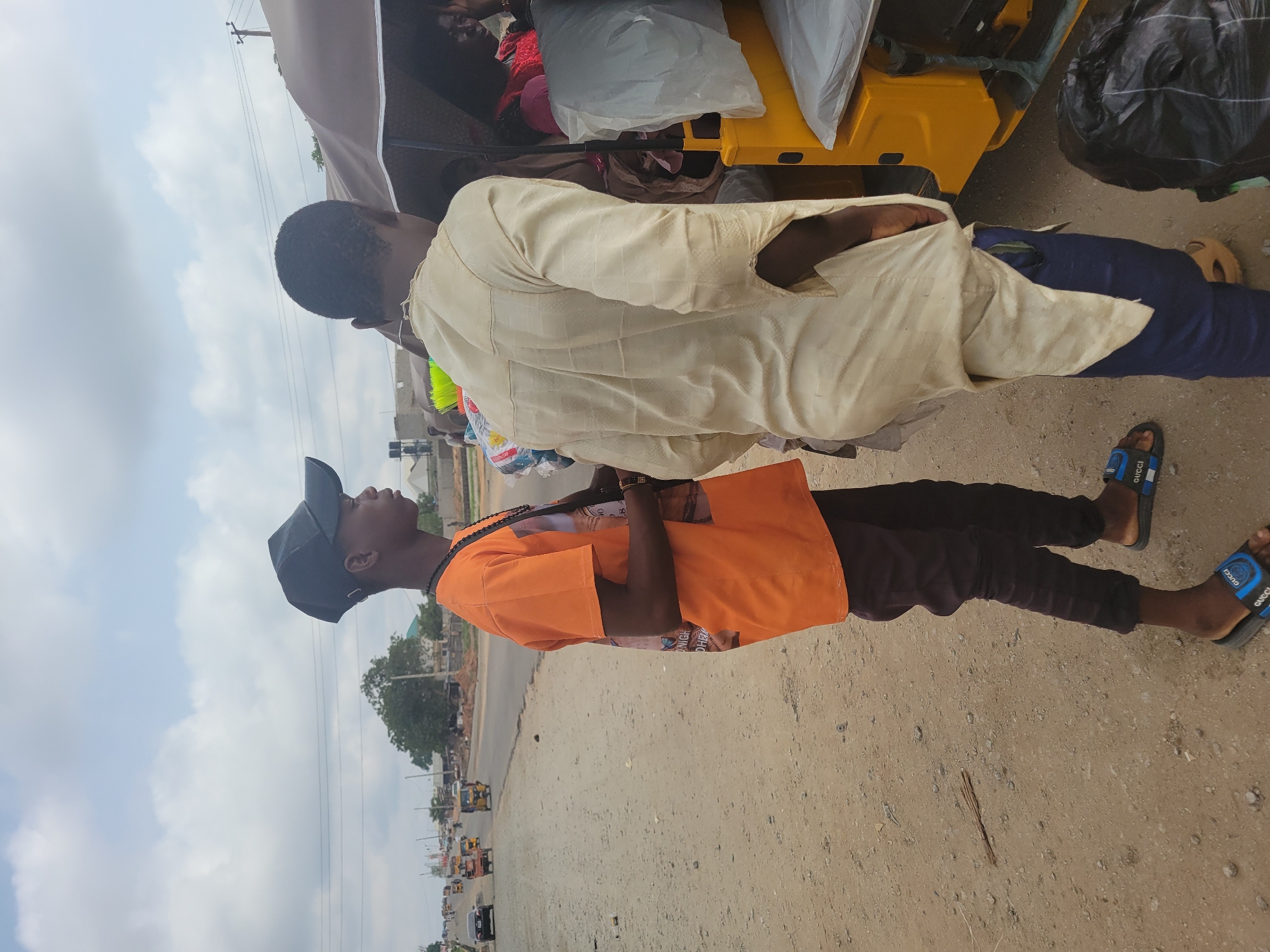 Two men are standing on a dirt shoulder of a road in Kwamba, Nigeria. One man is wearing a light yellow shirt and blue pants and has black sandals on his feet. The other man is wearing an orange shirt, dark-colored pants, and black and blue sandals. He has a black baseball cap on his head. They are standing next to a yellow vehicle that appears to be a commercial vehicle with a canvas top. There is baggage on top of the vehicle. The road has light traffic consisting of other commercial vehicles, cars, and motorcycles. The sky is overcast.