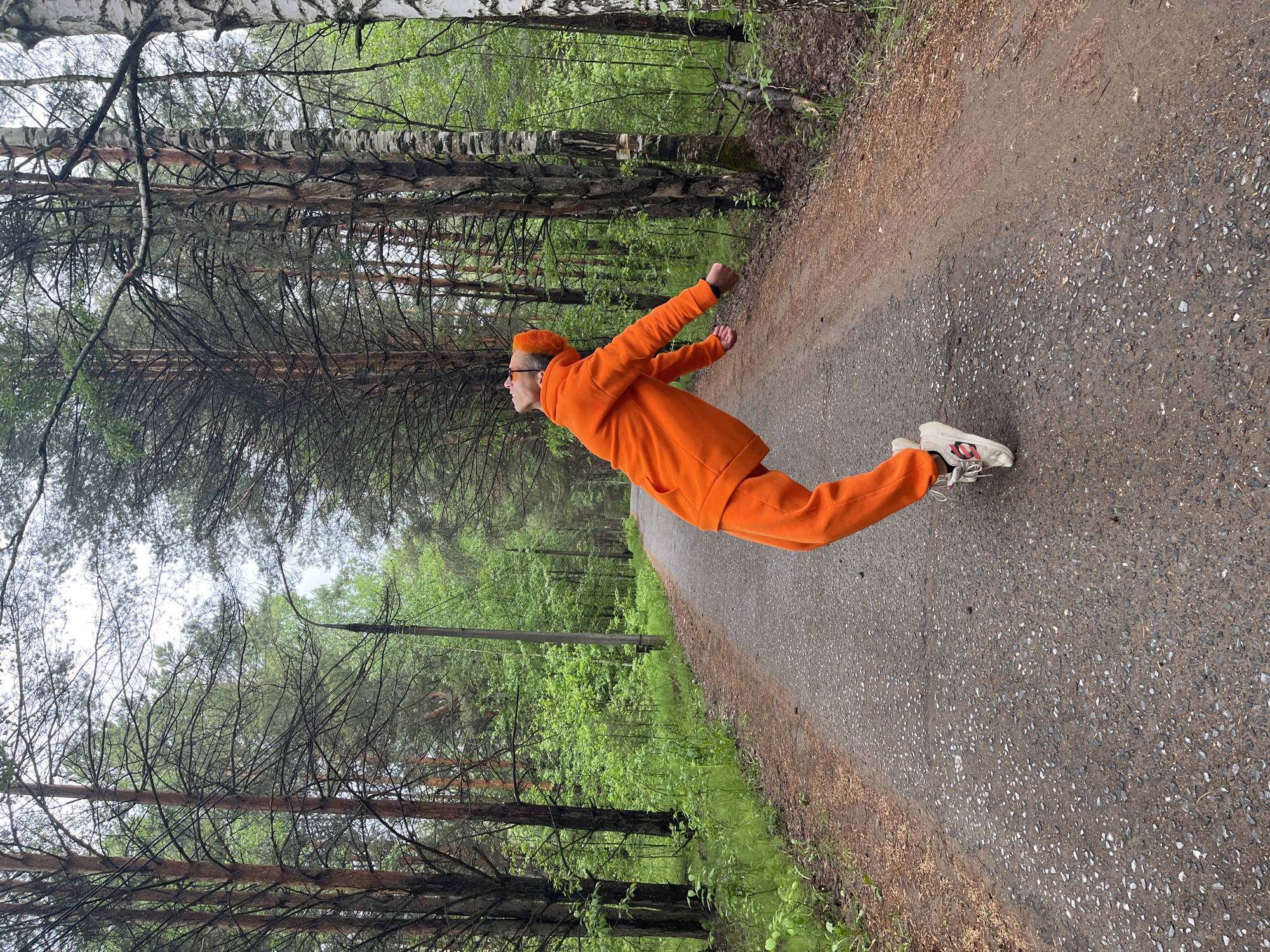 A person in an orange tracksuit lies horizontally on a pavement surrounded by trees. The person's body is mostly parallel to the edge of the pavement. The location appears to be a park or wooded area in the city of Chaikovsky, Russia.